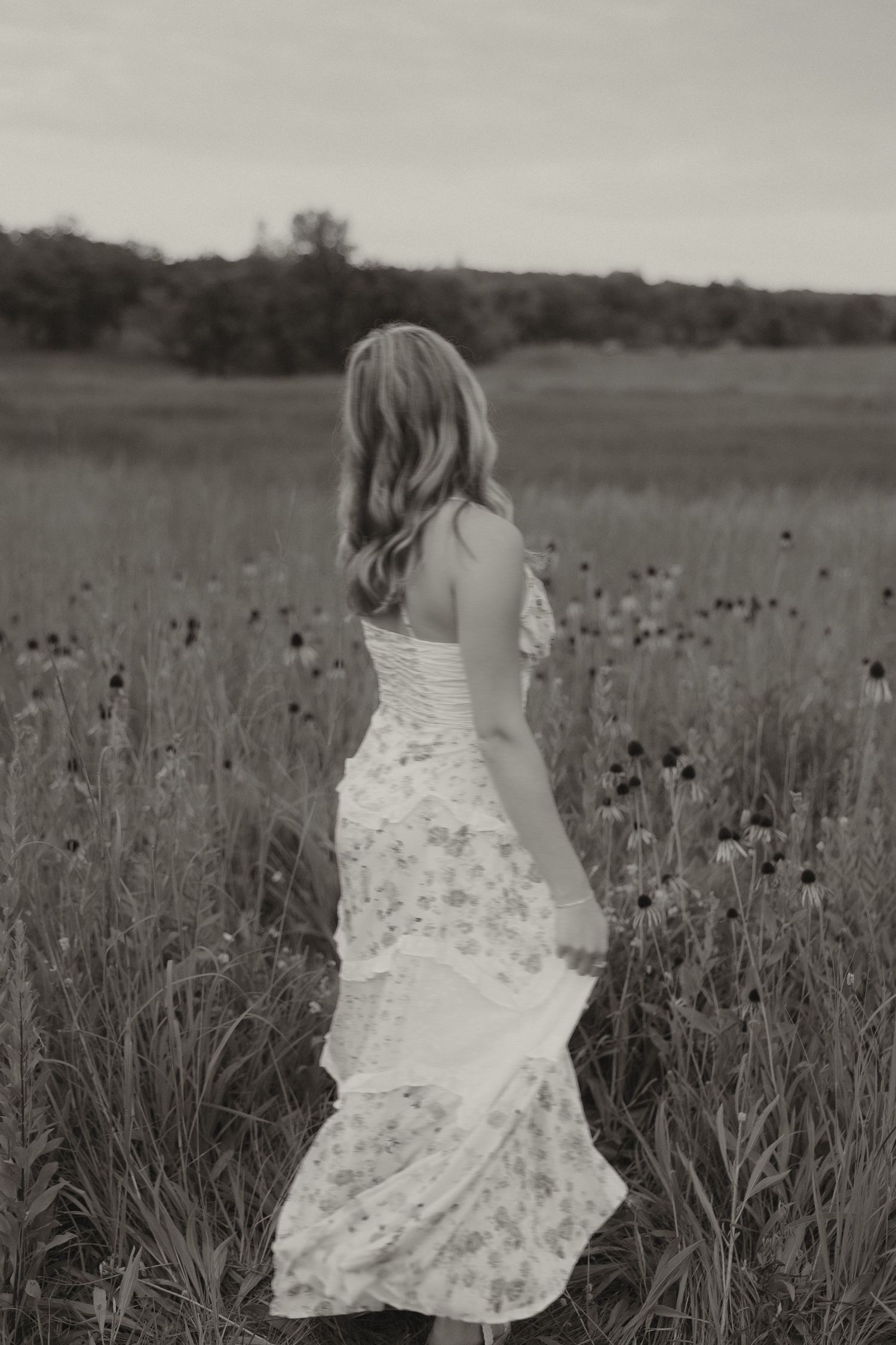 Black and white photo of blonde woman in floral dress walking through a wildflower meadow.