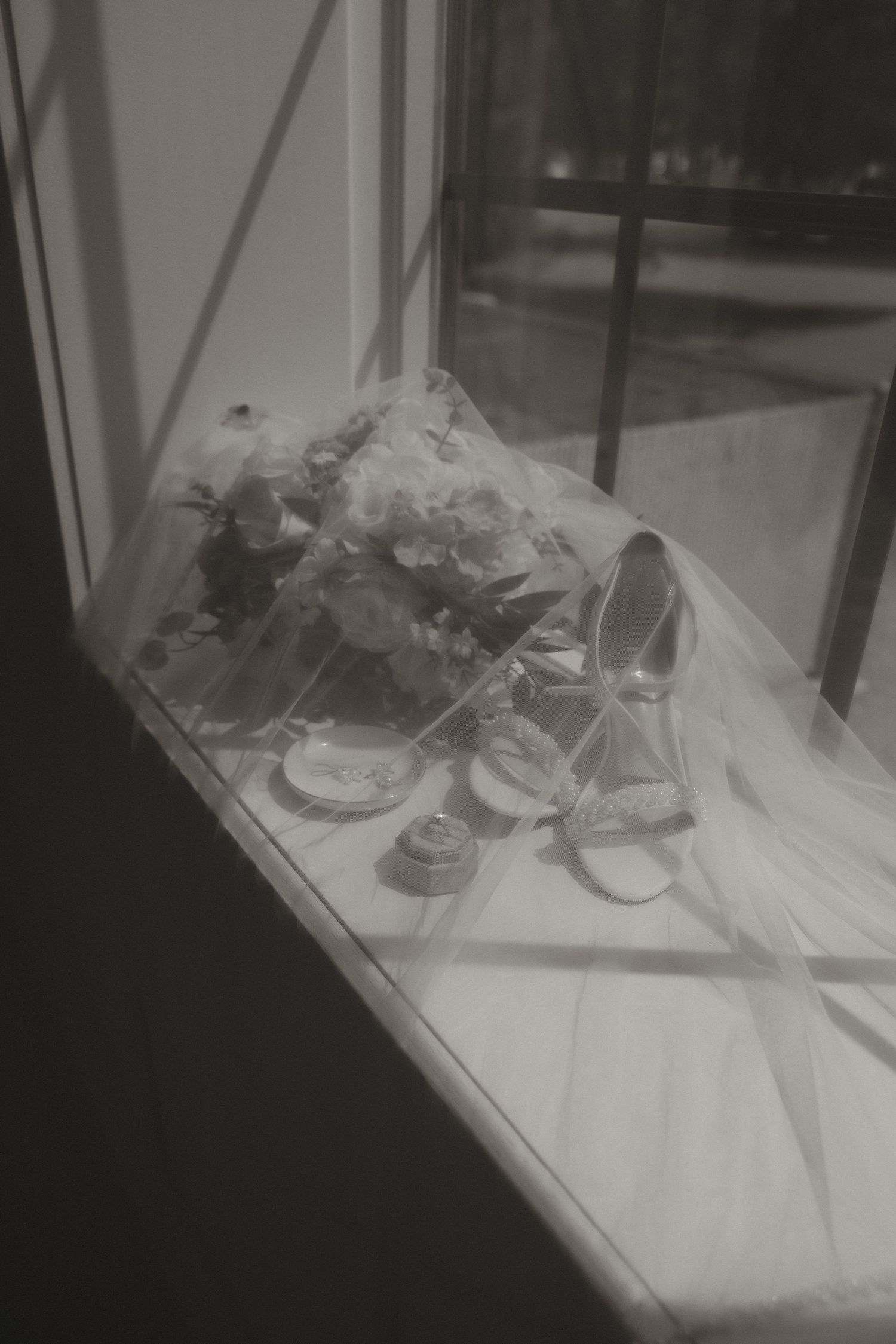 Black and white photo of bridal bouquet, heels, and accessories arranged on a sunlit windowsill.