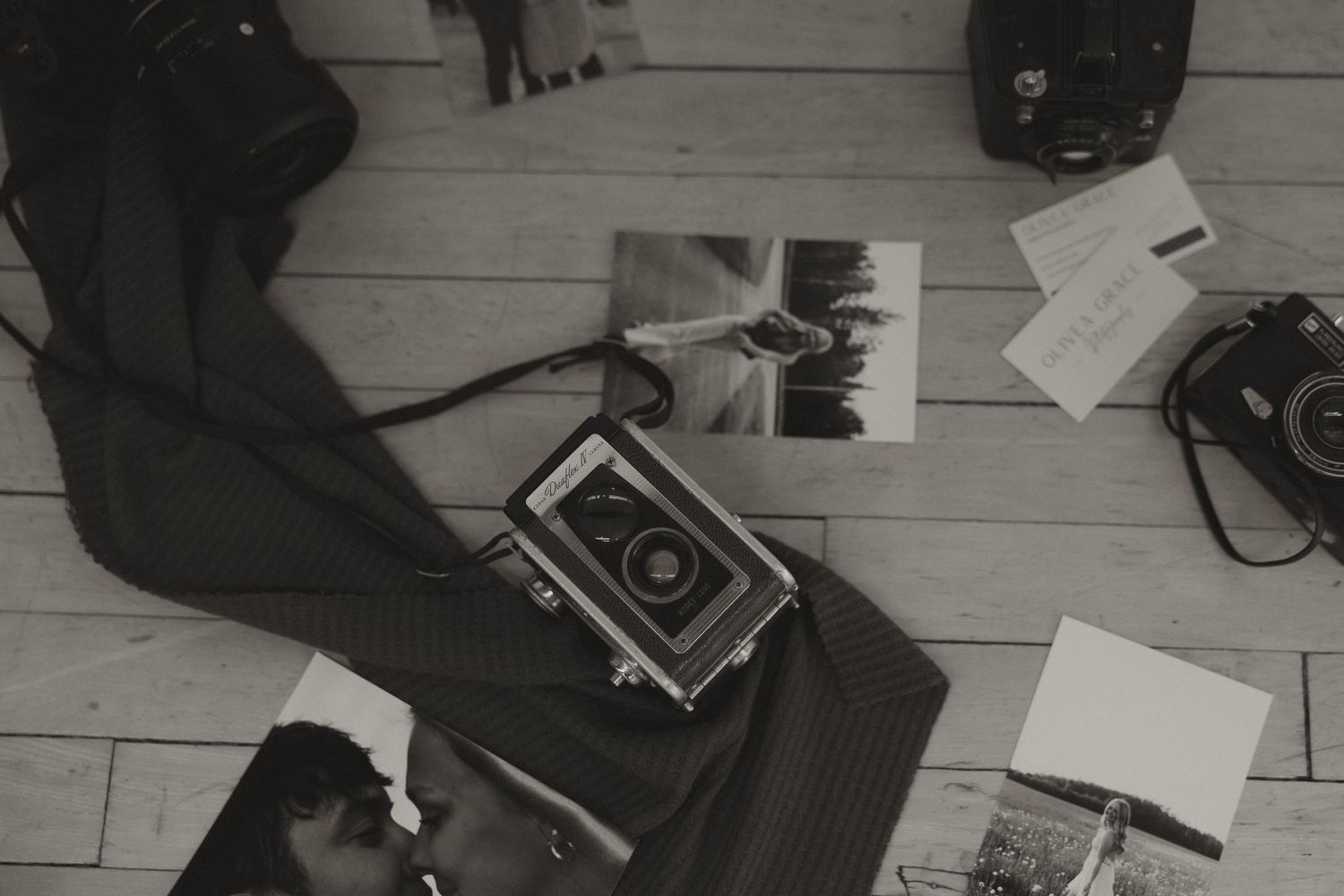 Black and white flat lay of vintage cameras, photos, and accessories on a wooden floor.