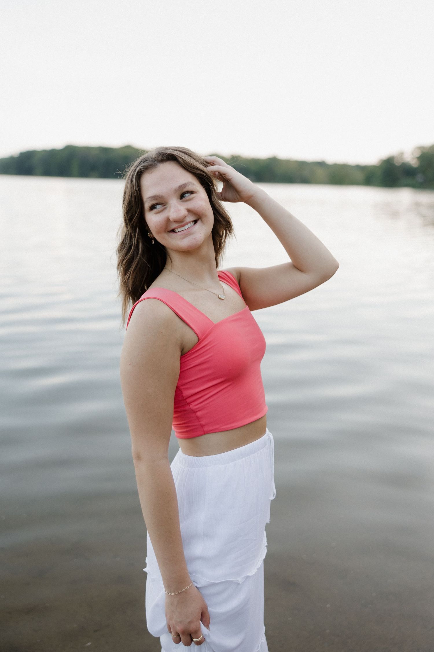 Smiling young woman in a coral crop top and white skirt standing by a calm lake at dusk.