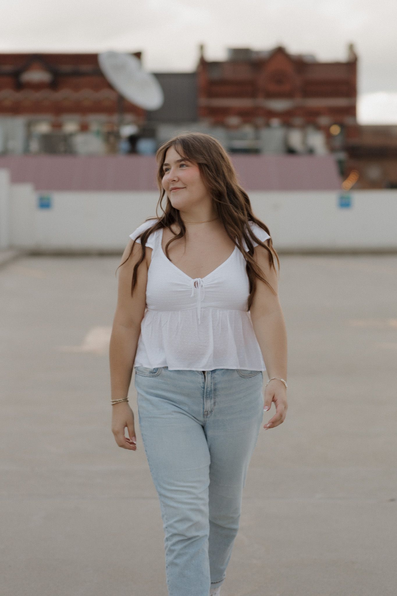 Smiling young woman in white top and jeans walking on rooftop with city buildings behind.