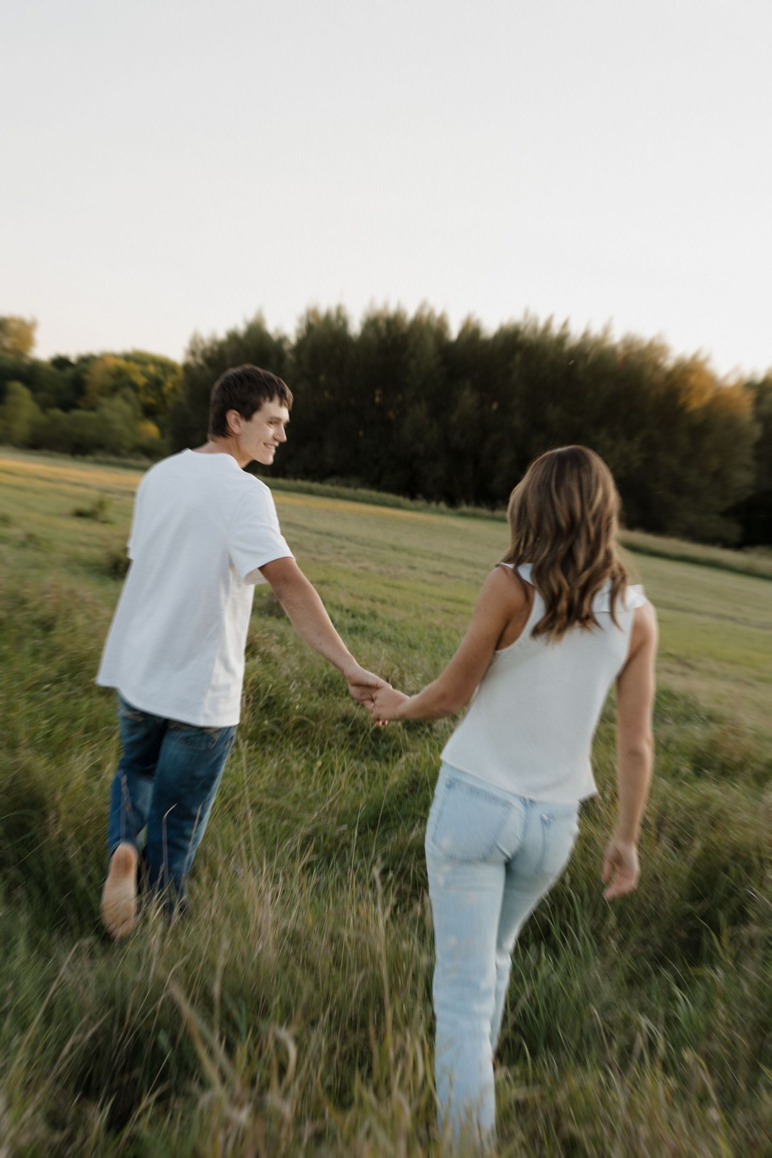 Couple holding hands and walking through a green meadow at sunset during engagement photo session.