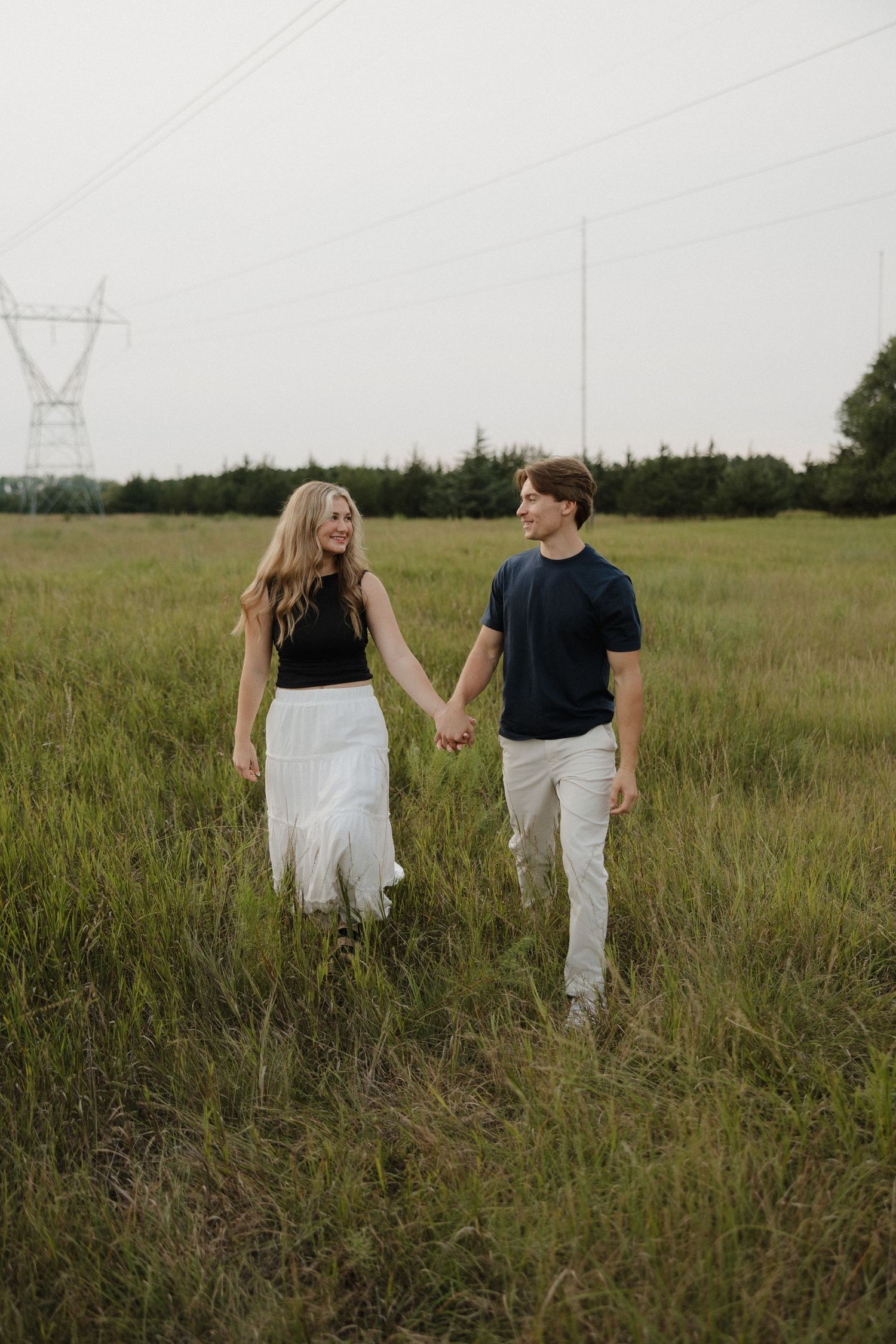 Happy couple holding hands and walking through a vast green field with power lines in background.