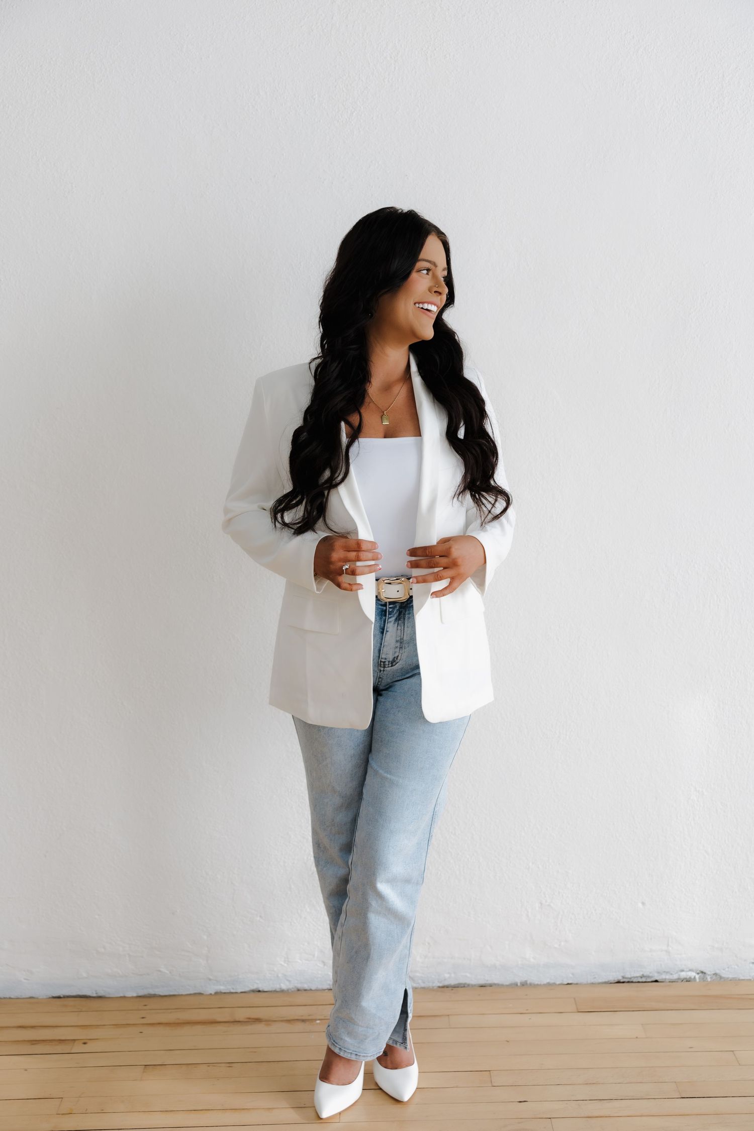 Smiling woman in white blazer, jeans, and white heels standing against a white wall holding a glass.