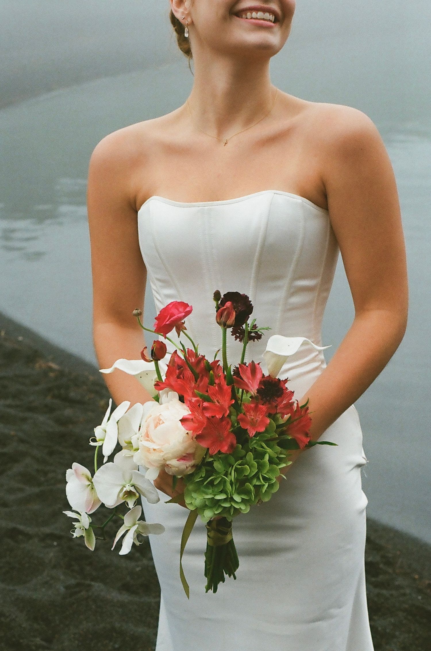 Smiling bride in strapless white gown holding vibrant bouquet of red, white, and green flowers outdoors.