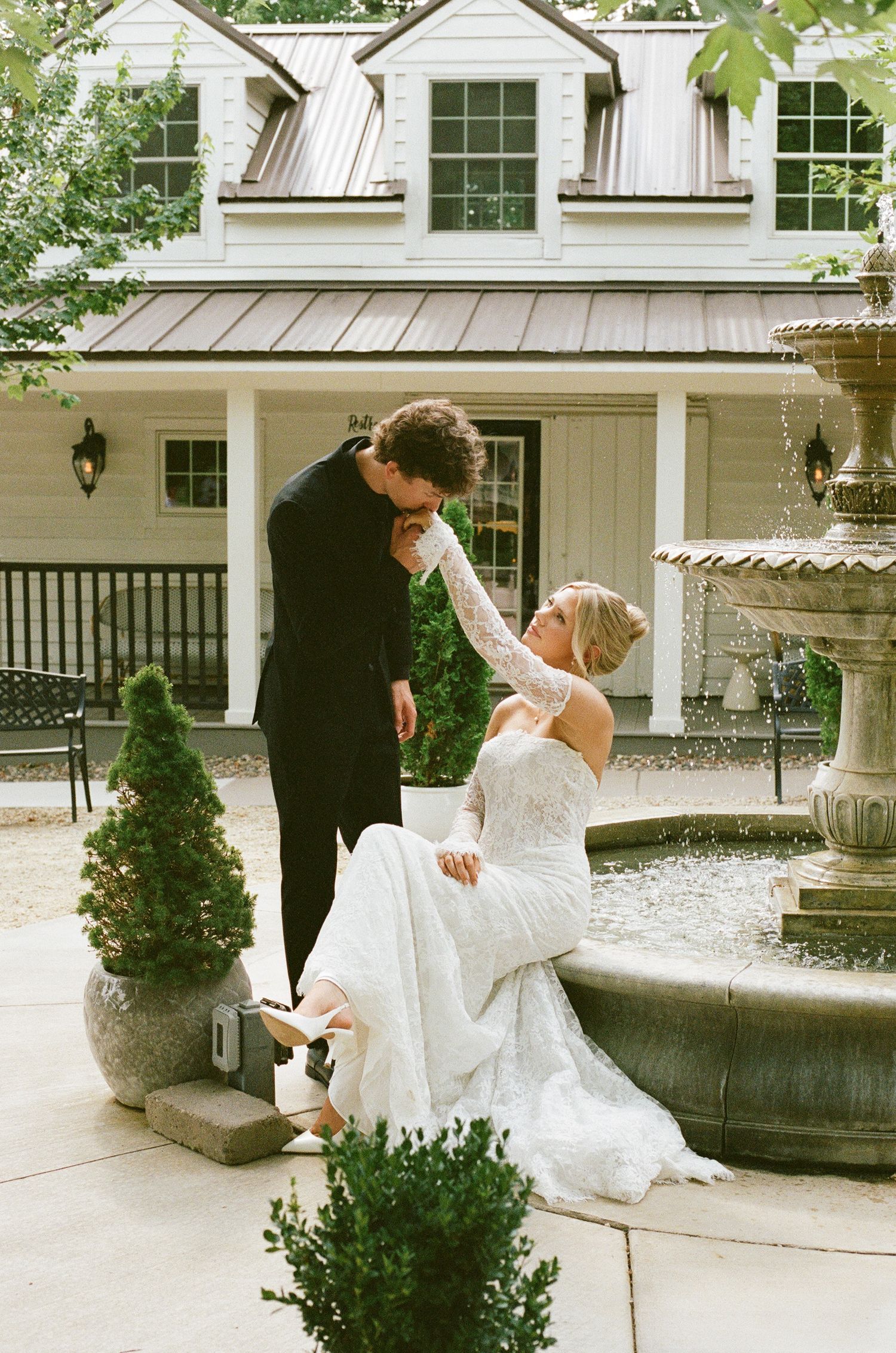 Groom in black suit kisses bride's hand as she sits by stone fountain outside white colonial wedding venue.