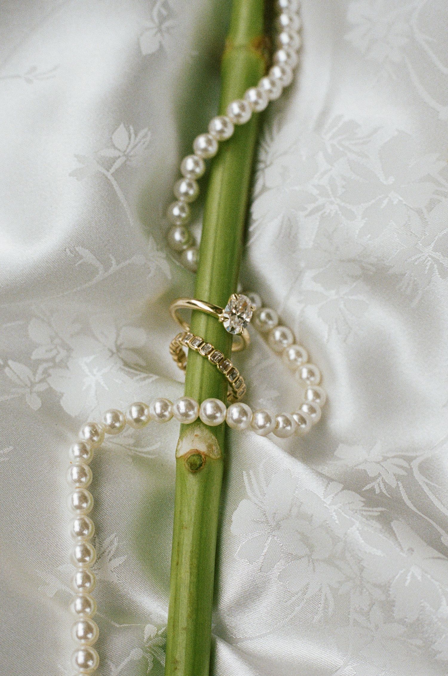 Pearl necklace and diamond ring draped over a green bamboo stem on white satin fabric.