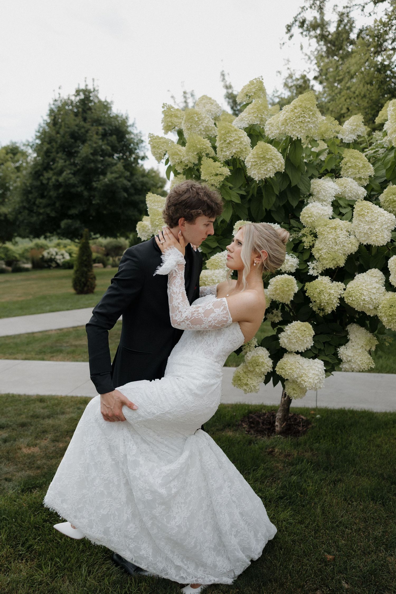 A bride and groom share a romantic dip pose in front of lush white hydrangea blooms in a garden setting.