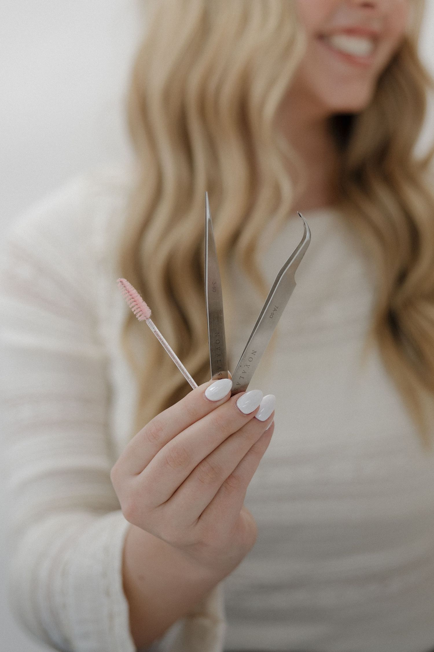 Smiling blonde woman holding lash tweezers, scissors, and a spoolie brush with white manicured nails.