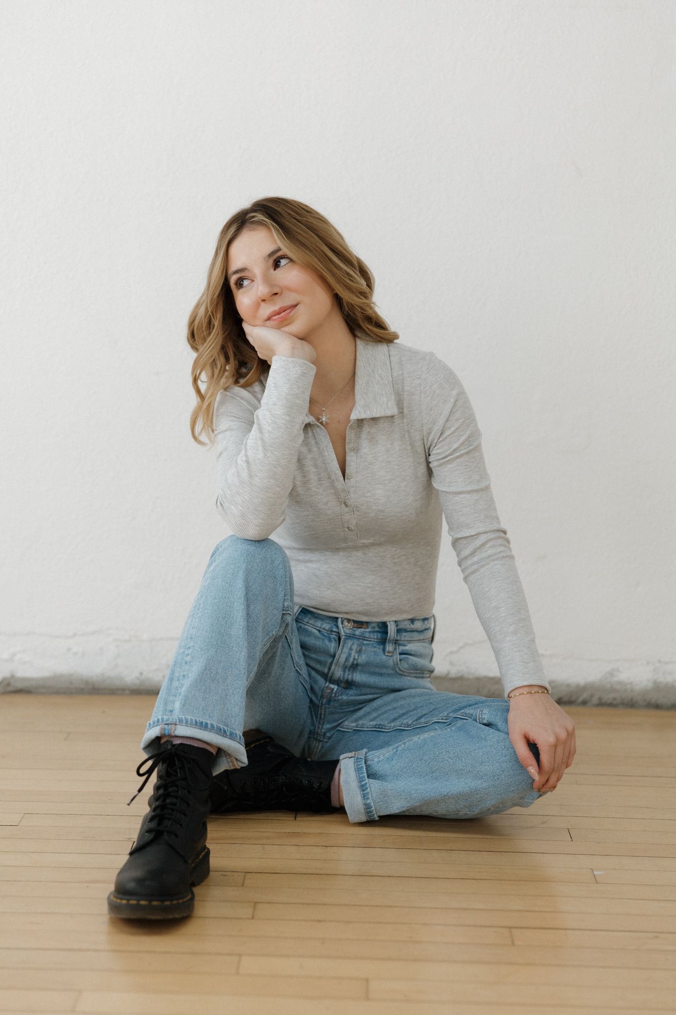 Smiling young woman in gray polo and jeans sitting on a hardwood floor against a white wall.