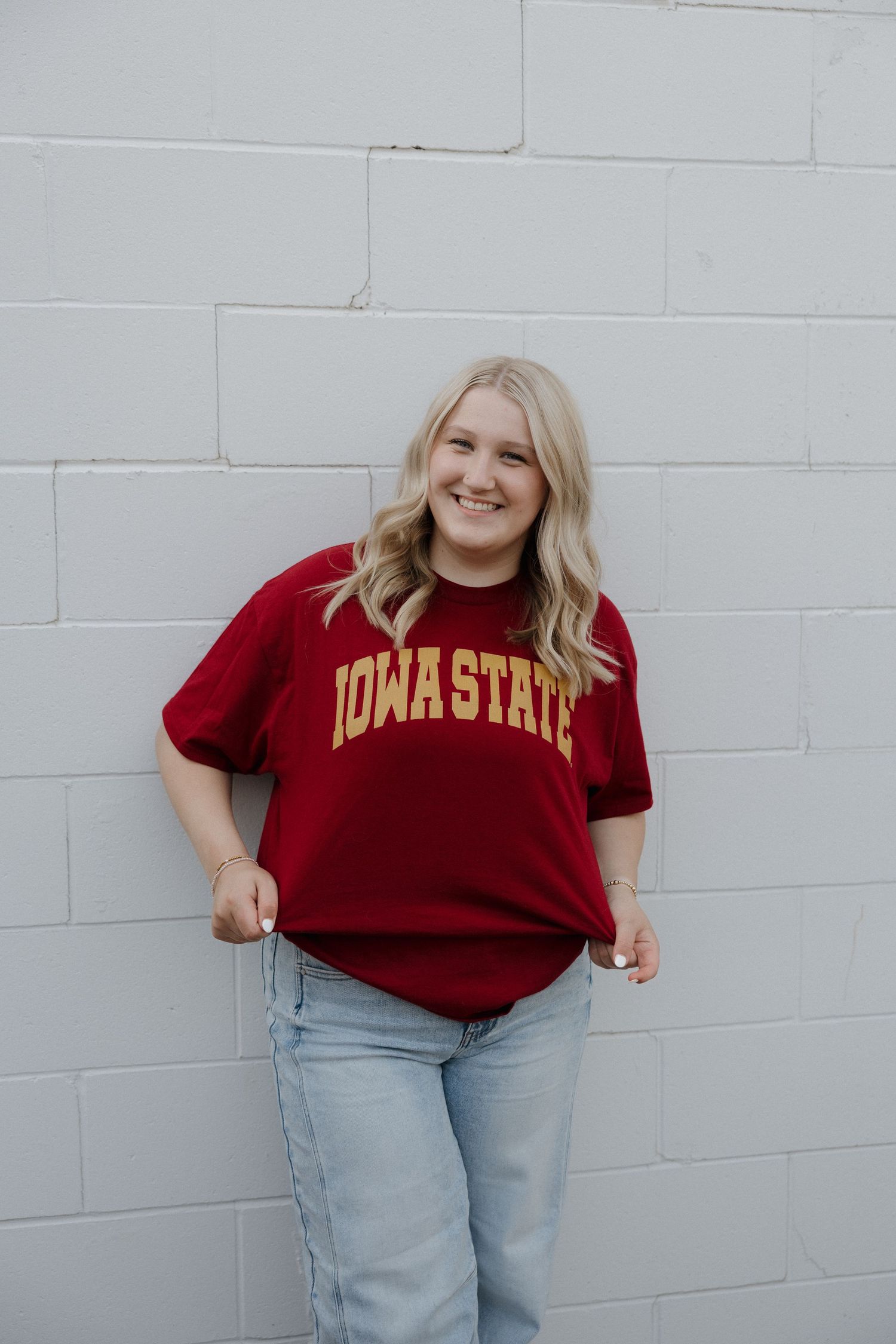 Smiling blonde woman wearing red Iowa State t-shirt leaning on white brick wall.