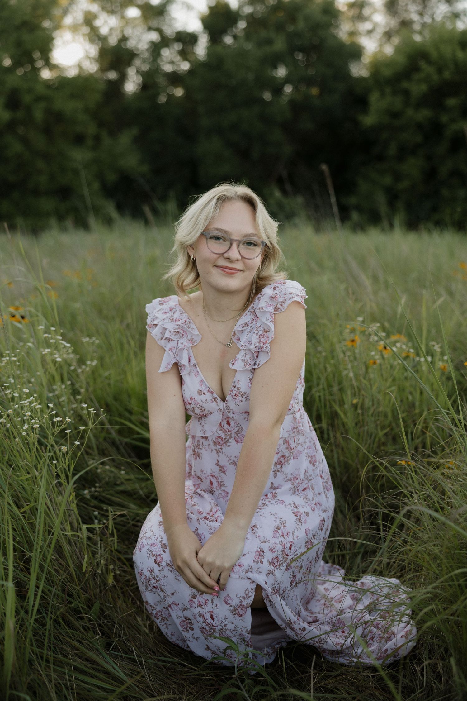 Smiling blonde woman with glasses in floral dress sitting in lush green meadow.