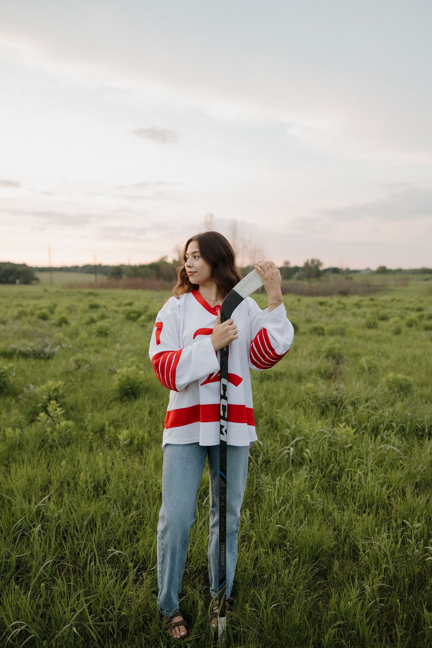 Young woman in hockey jersey holding a hockey stick upright while standing in a green field at dusk.