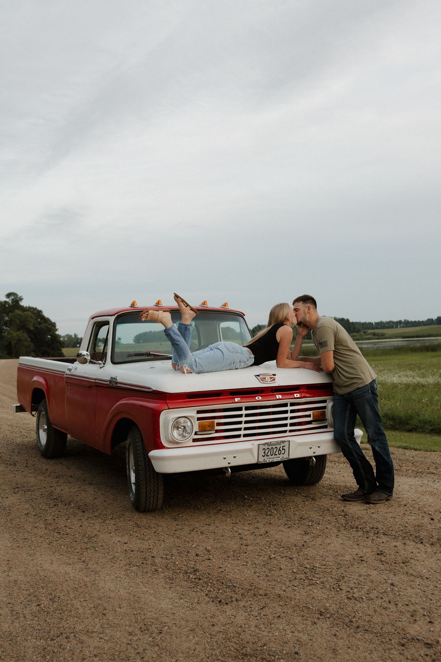 Woman lying on vintage red Ford truck hood while couple kisses on dirt road.