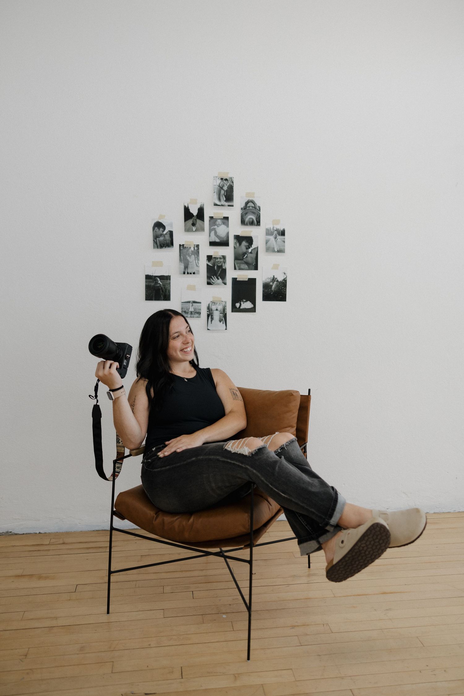 Woman sitting in brown leather chair holding a camera, with black and white photos displayed on wall behind her.