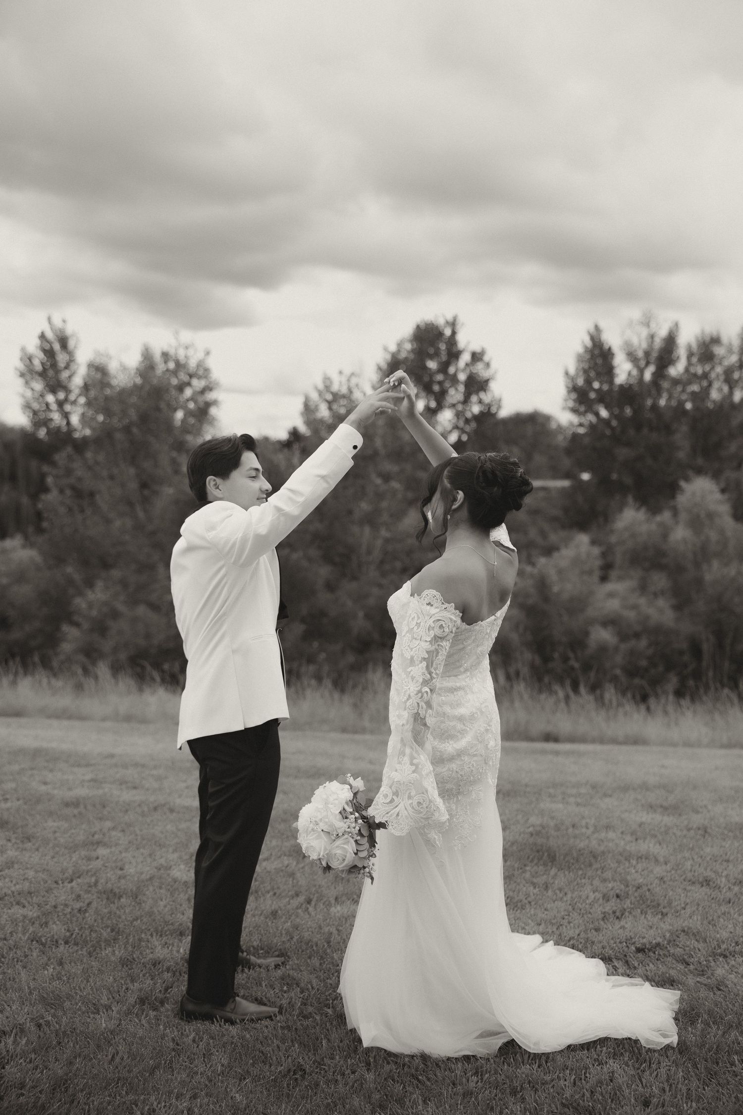 Black and white photo of a couple in wedding attire posing in a field under cloudy skies.