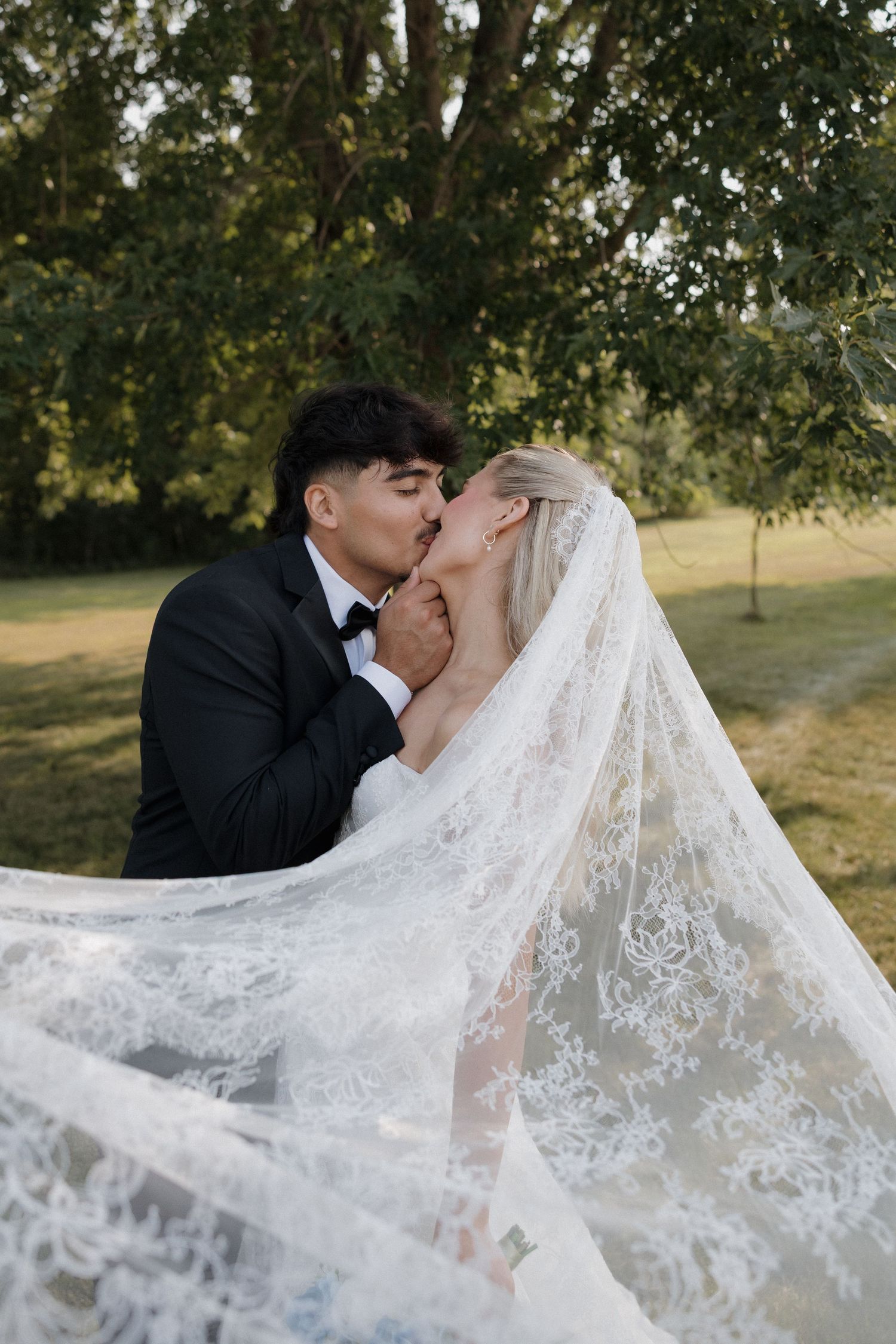 Bride and groom share a romantic kiss outdoors, with flowing lace wedding veil, surrounded by lush green trees.