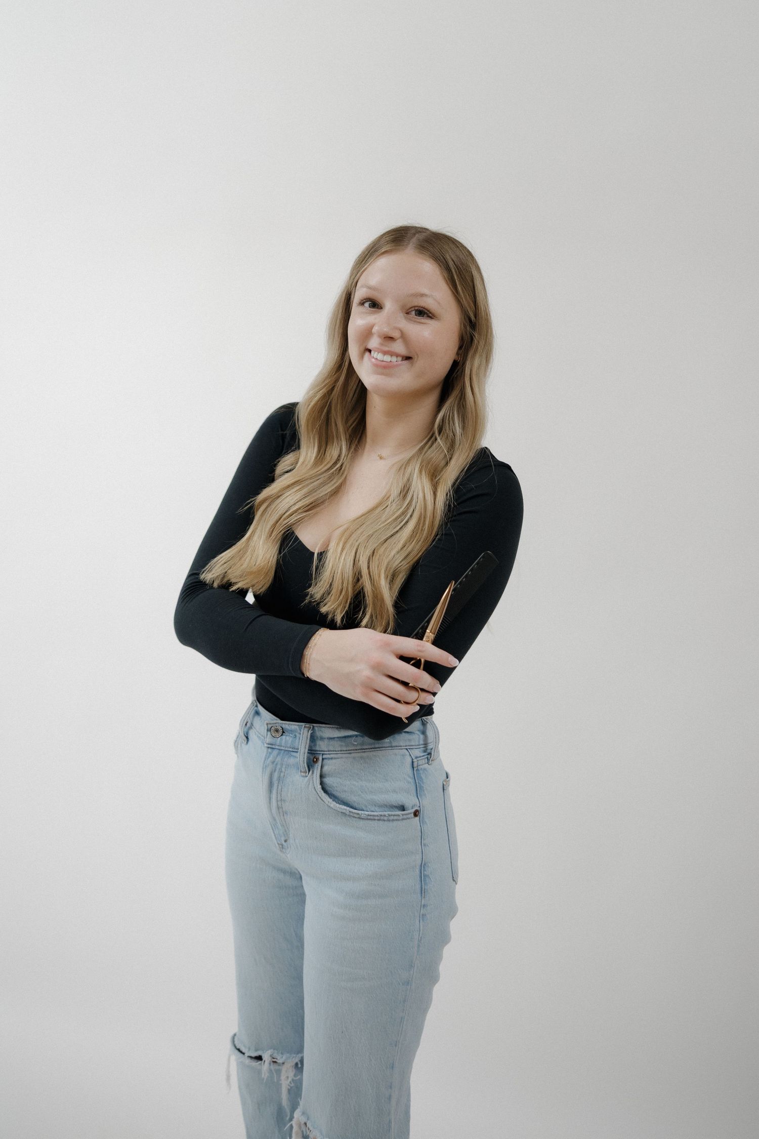 Smiling young blonde woman in black top and ripped jeans holding a hair brush against white background.