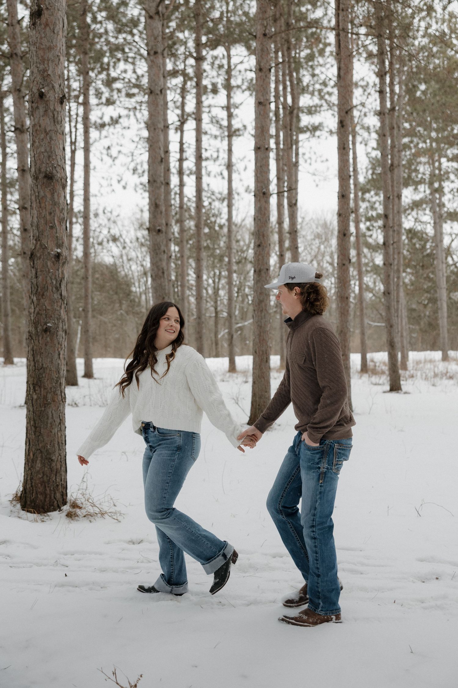 Happy couple holding hands and walking through a snowy pine forest, woman in white sweater and jeans smiling.