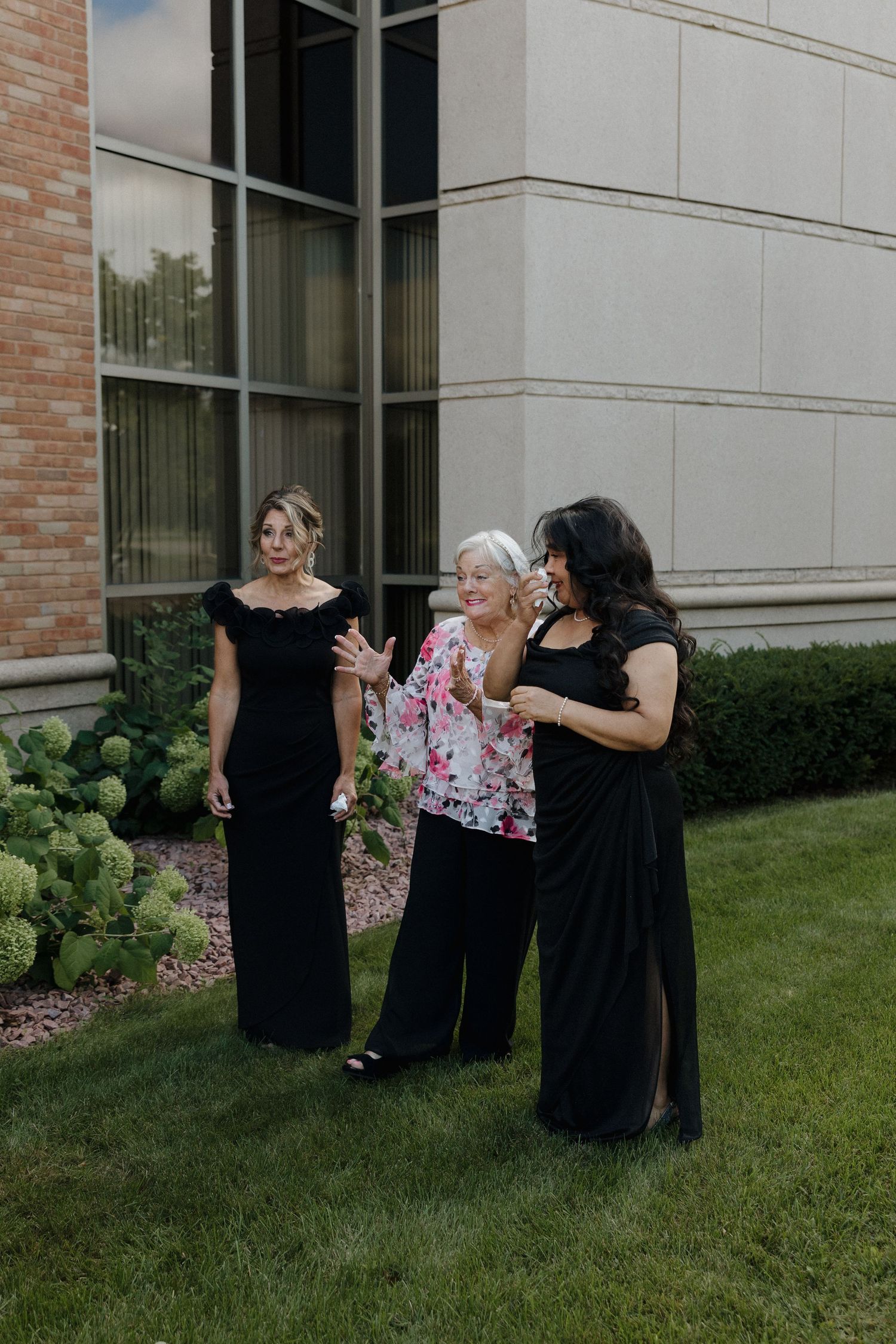 Wedding party members in black dresses share embraces on a grassy lawn.
