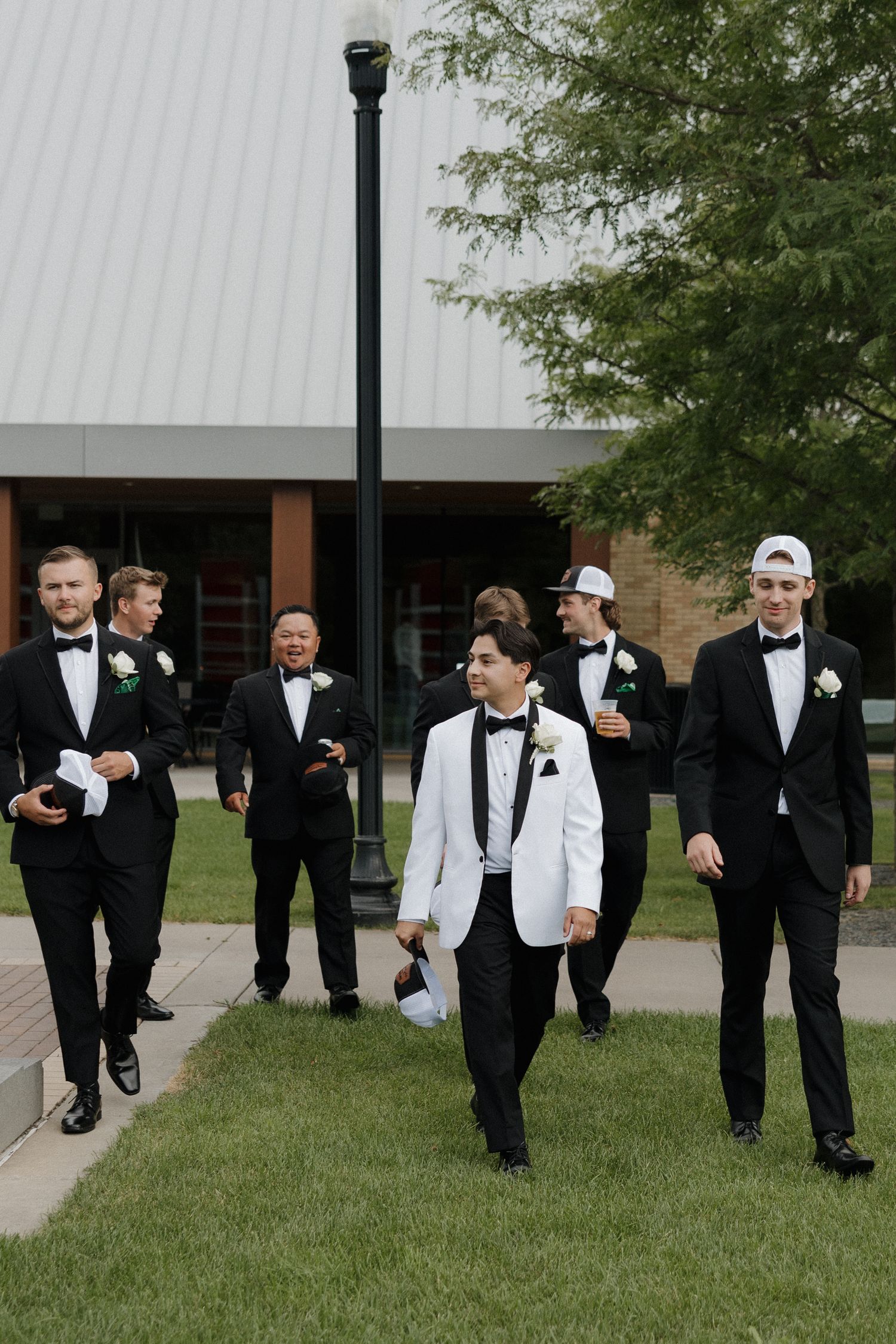Groomsmen in black tuxedos and white shirts walking together on grass outside a building.