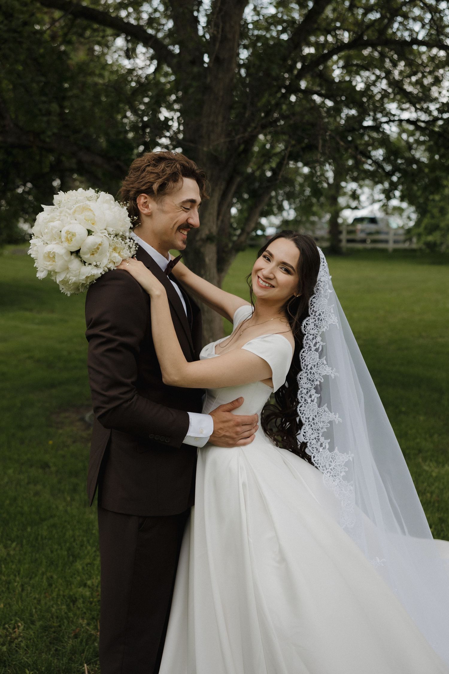 A romantic wedding portrait shows a couple embracing outdoors with a white bouquet and flowing lace veil.