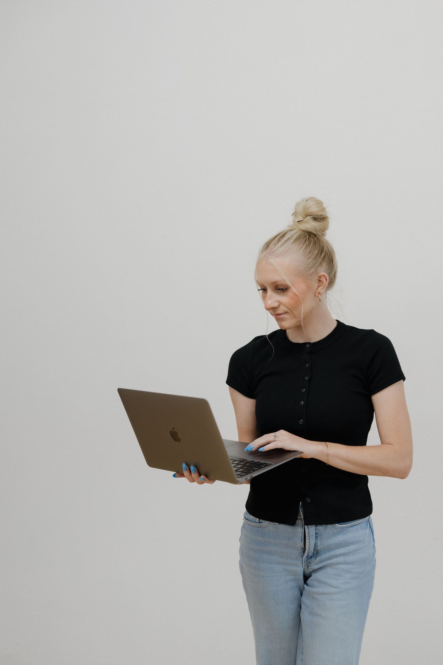 Blonde woman in black top and jeans standing while using a laptop against white background.