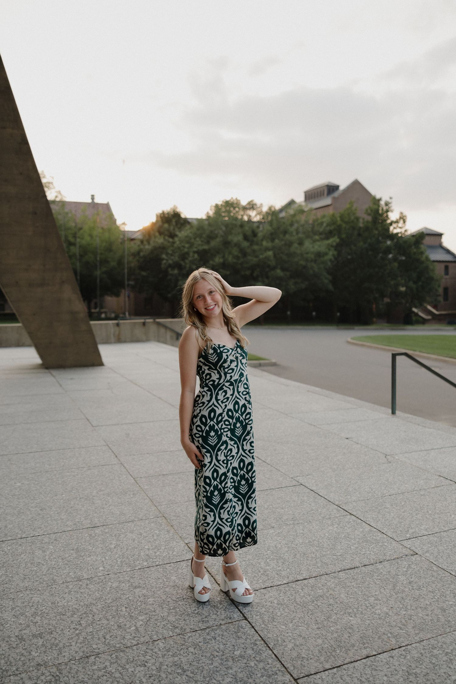 Smiling blonde woman in green patterned midi dress posing on tiled plaza with trees and buildings in background.