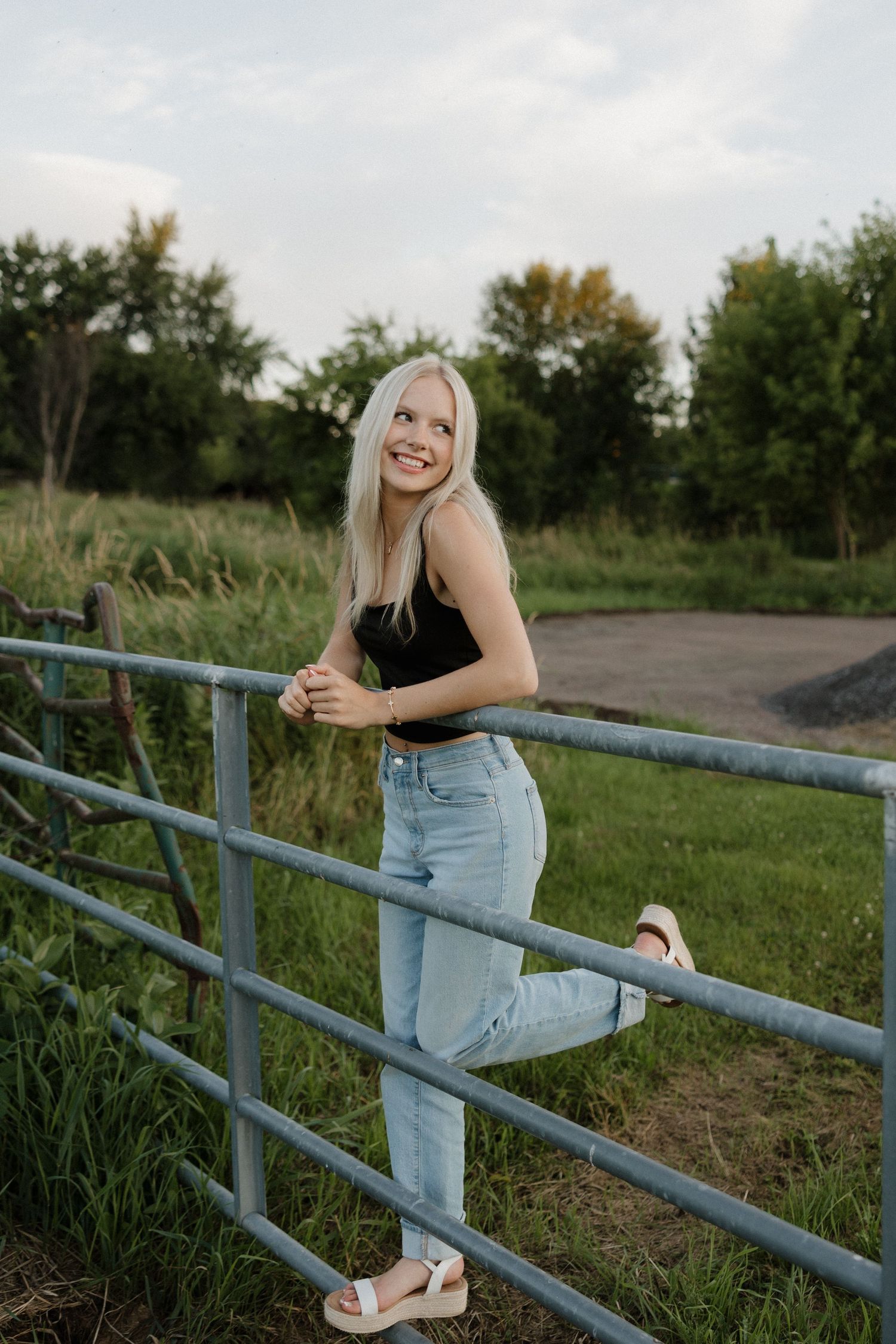 Smiling blonde woman in black tank top and jeans leaning on a metal farm gate in a green rural setting.
