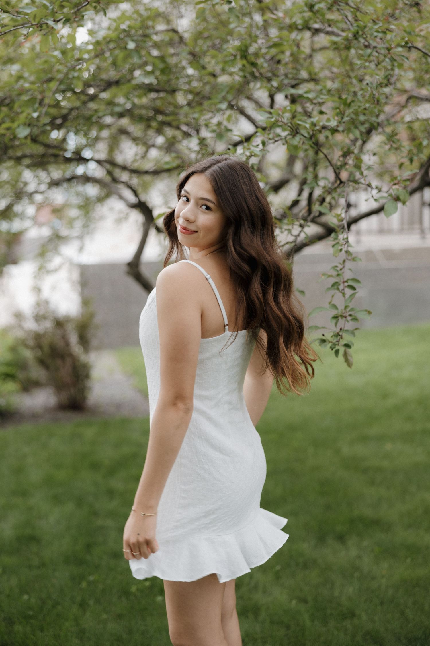 Young woman in white mini dress standing under blooming tree in garden.