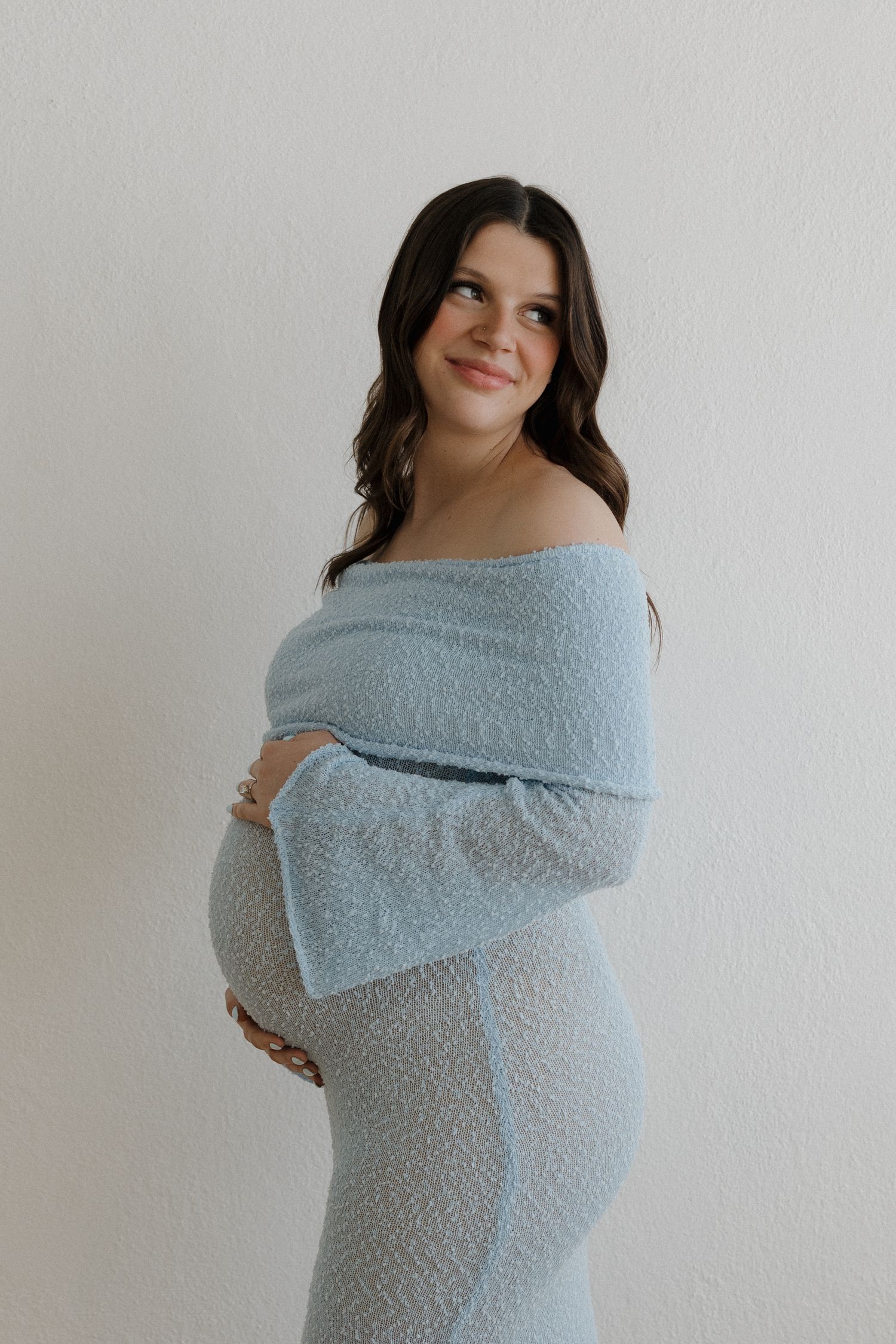Pregnant woman in light blue off-shoulder dress smiling softly, cradling her baby bump against white background.