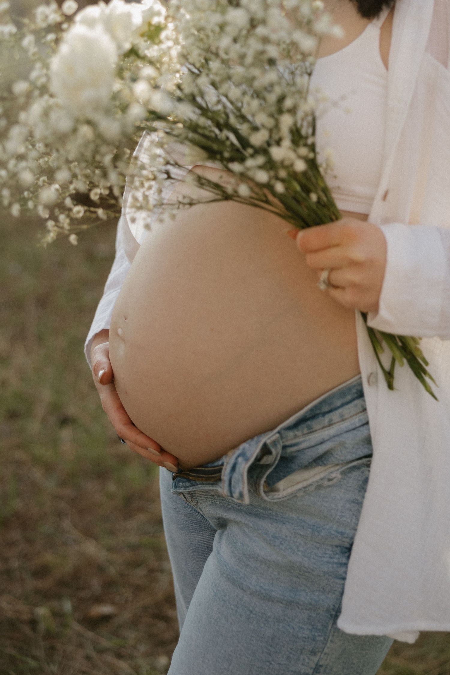 Close-up of pregnant belly with white flower bouquet, woman in open white shirt and blue jeans outdoors.