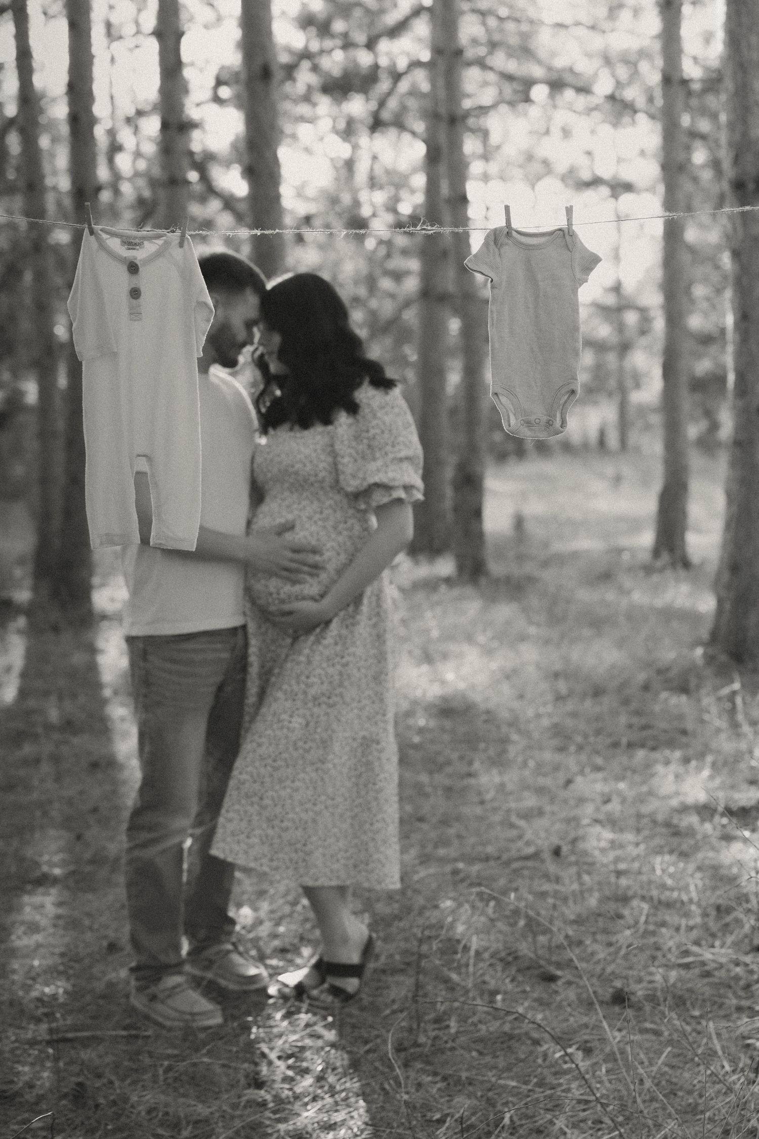 Black and white photo of pregnant couple embracing in forest with baby clothes hanging on a line.
