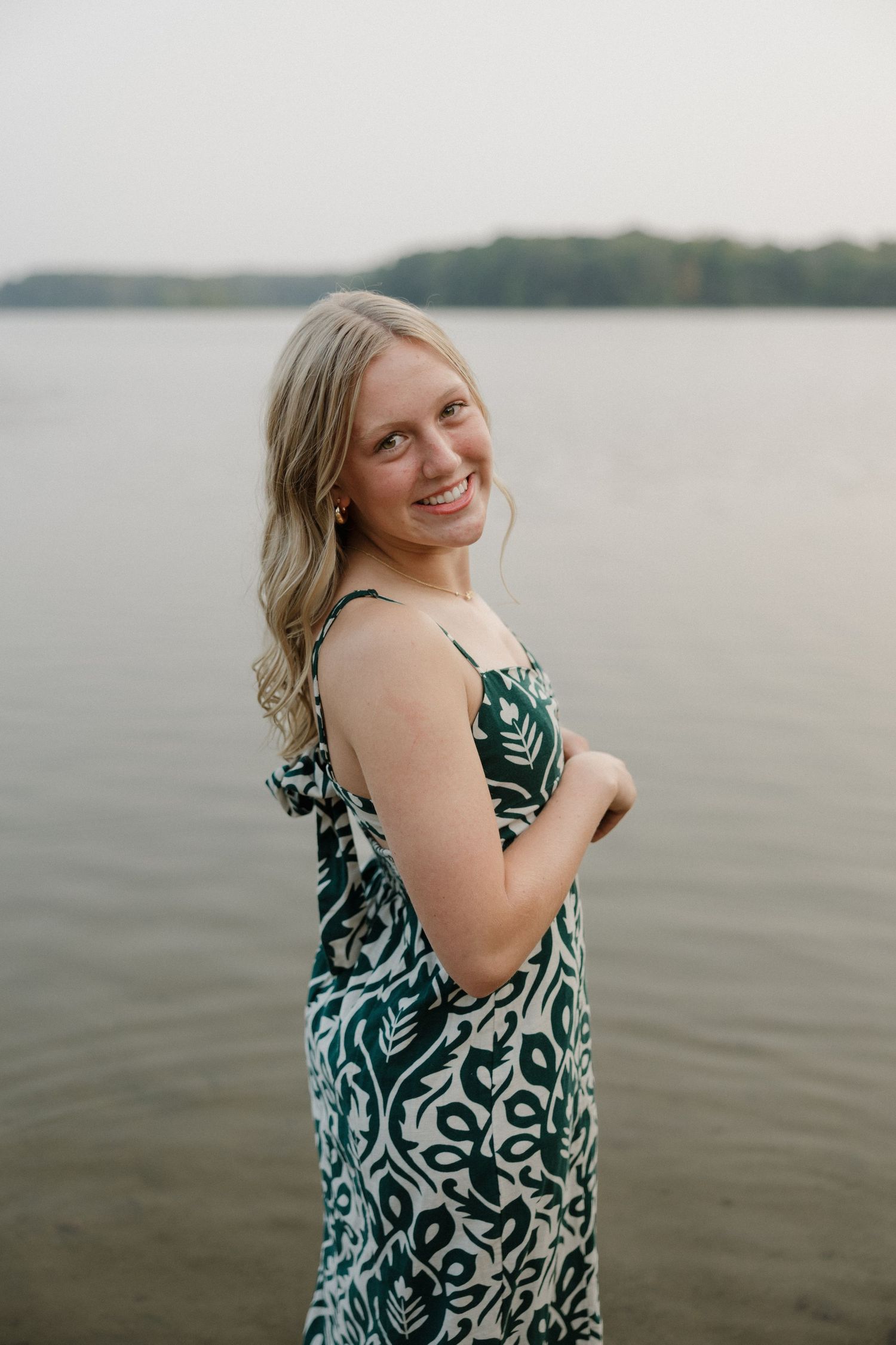 Smiling blonde woman in green floral patterned dress standing in calm lake water with treeline in background.