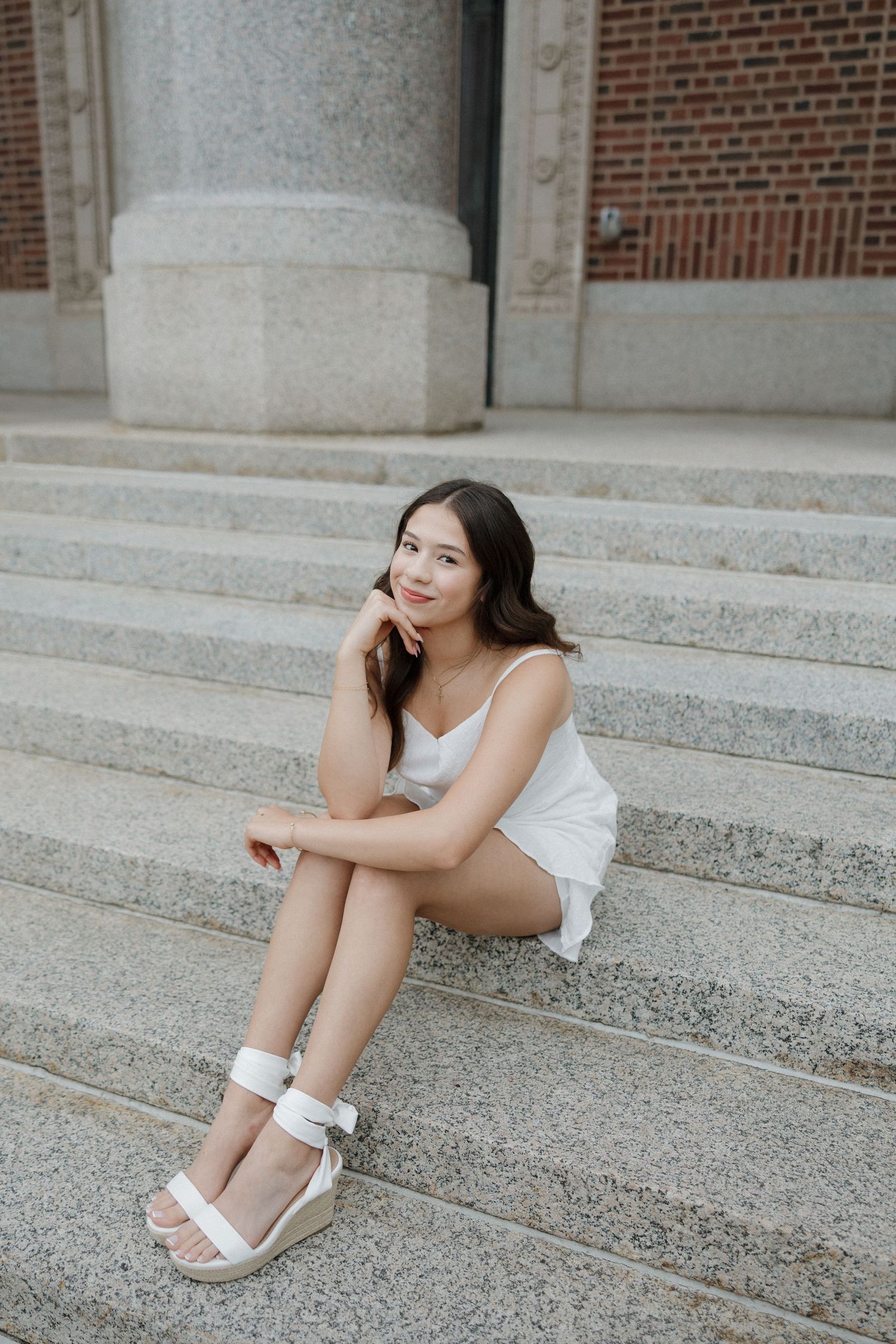 Young woman in white dress sitting on stone steps by brick building.