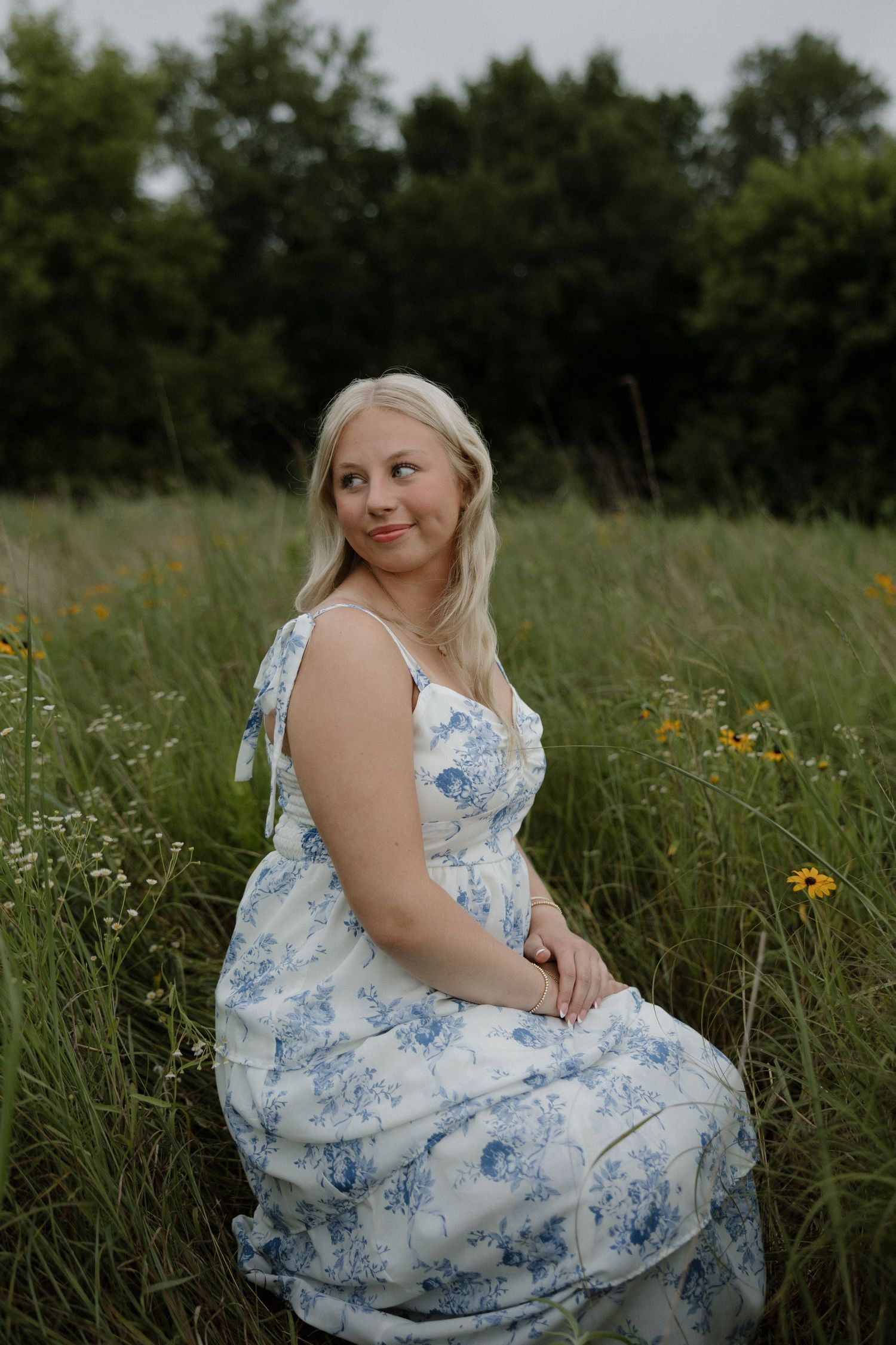 Blonde woman in a white and blue floral dress sitting gracefully in a lush green wildflower meadow.