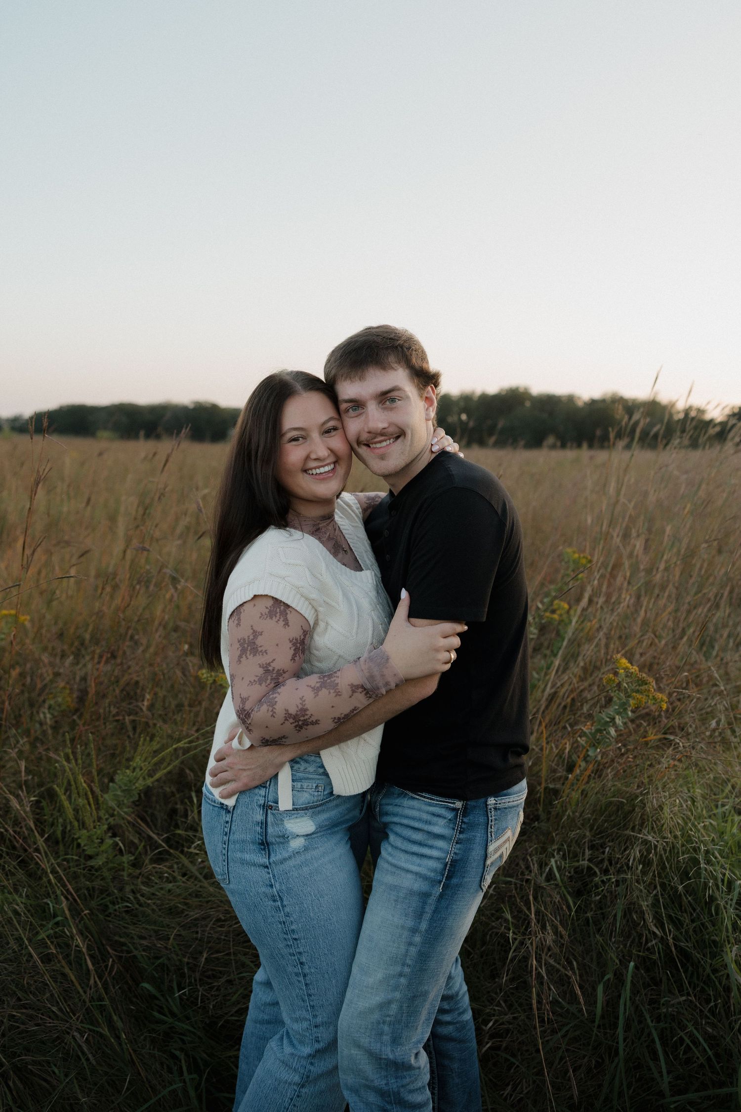 Smiling couple hugging in a golden field at dusk, woman showing floral arm tattoos.