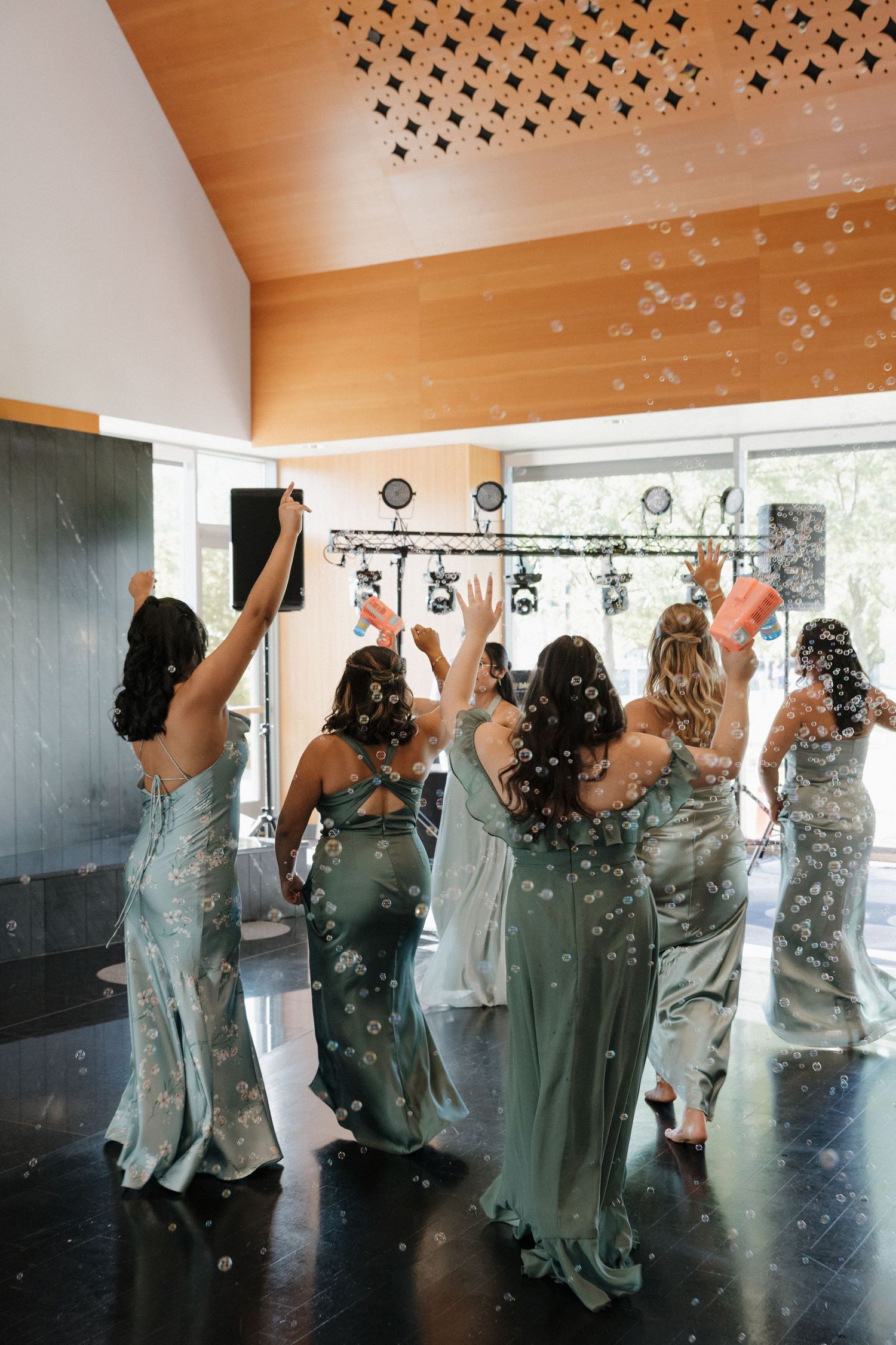 Wedding party dancing and celebrating with bubbles floating through the air in an indoor venue.