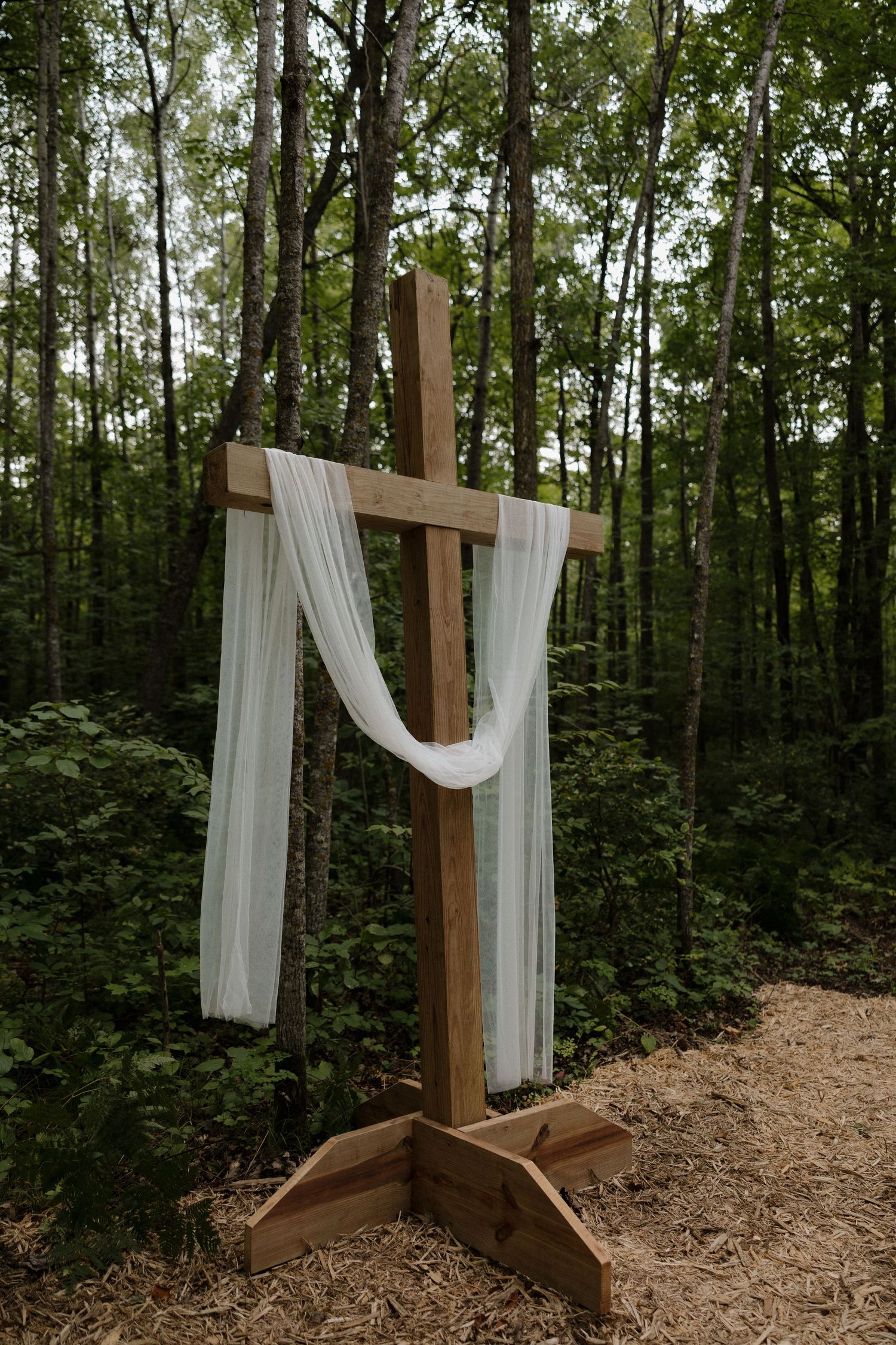 Wooden crosses draped with flowing white fabric create a rustic wedding altar in a wooded forest setting.