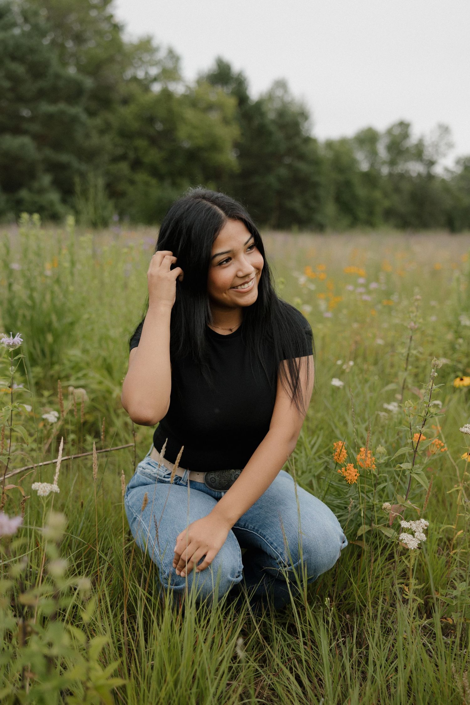 Young woman with black hair in black tee and jeans kneeling in colorful wildflower meadow smiling.