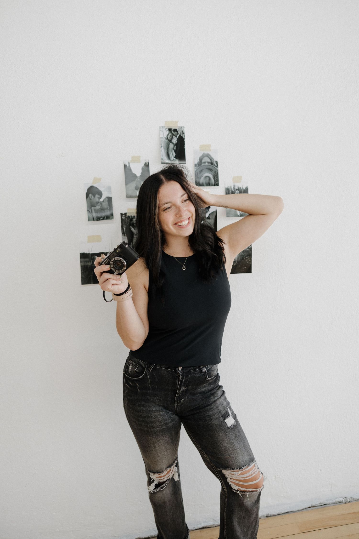 Smiling brunette woman in black tank top holding camera, leaning against wall with black and white photos.