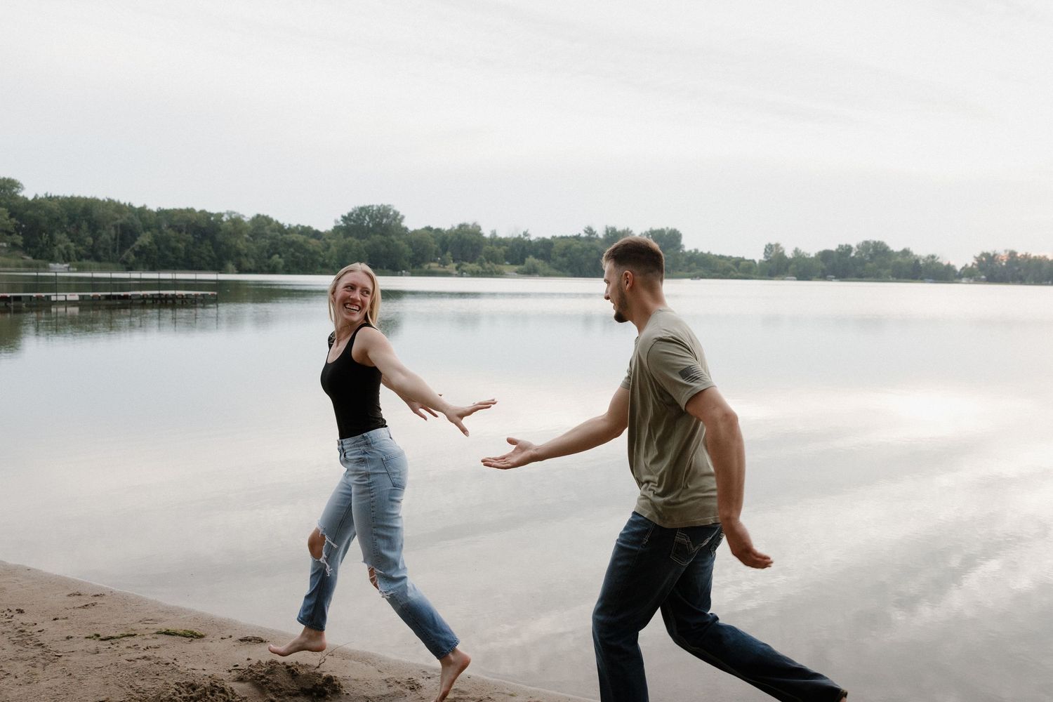 Laughing blonde woman reaching toward man walking along a lakeside beach.