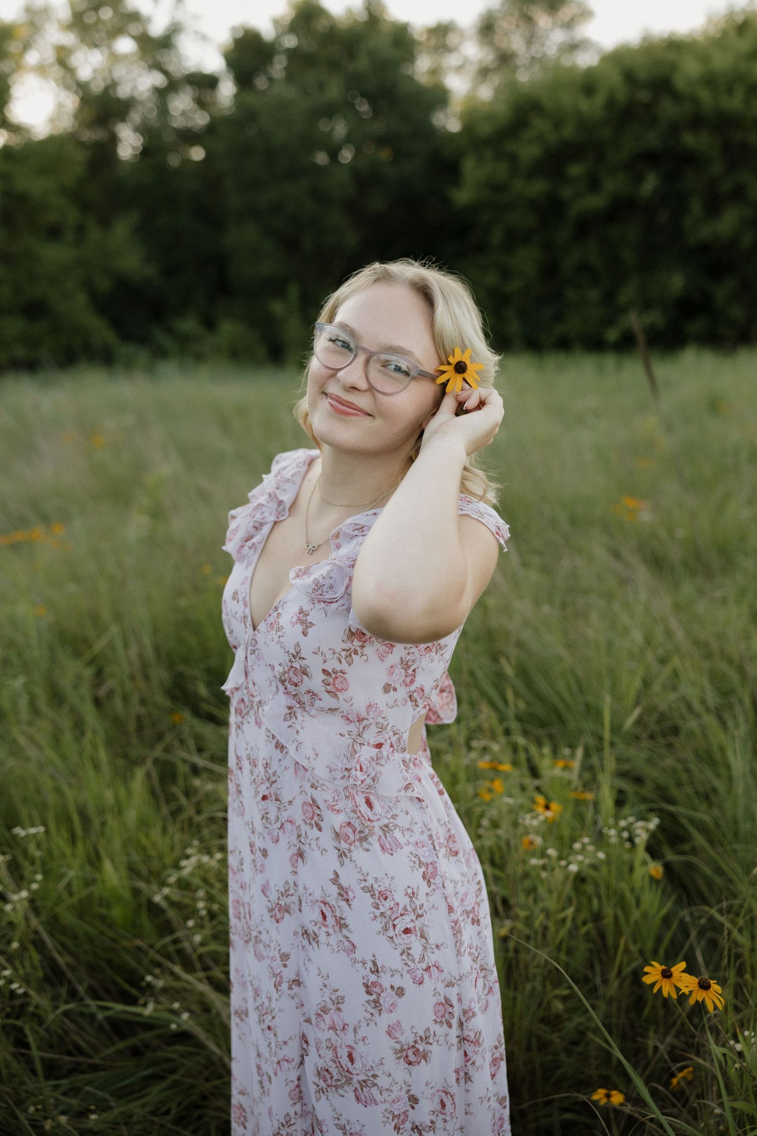 Blonde woman with glasses holding yellow flower near ear in green meadow field.