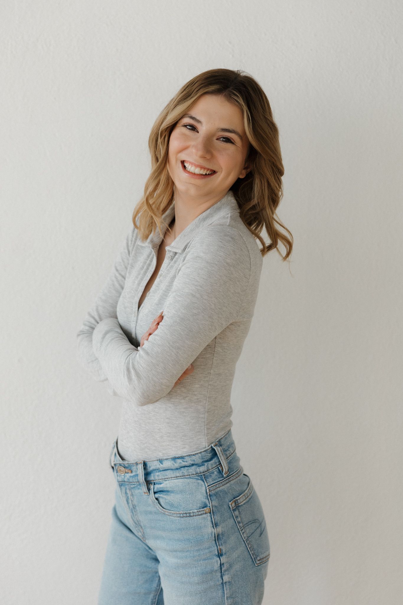 Smiling young woman in gray polo and jeans posing with arms crossed against a white wall.