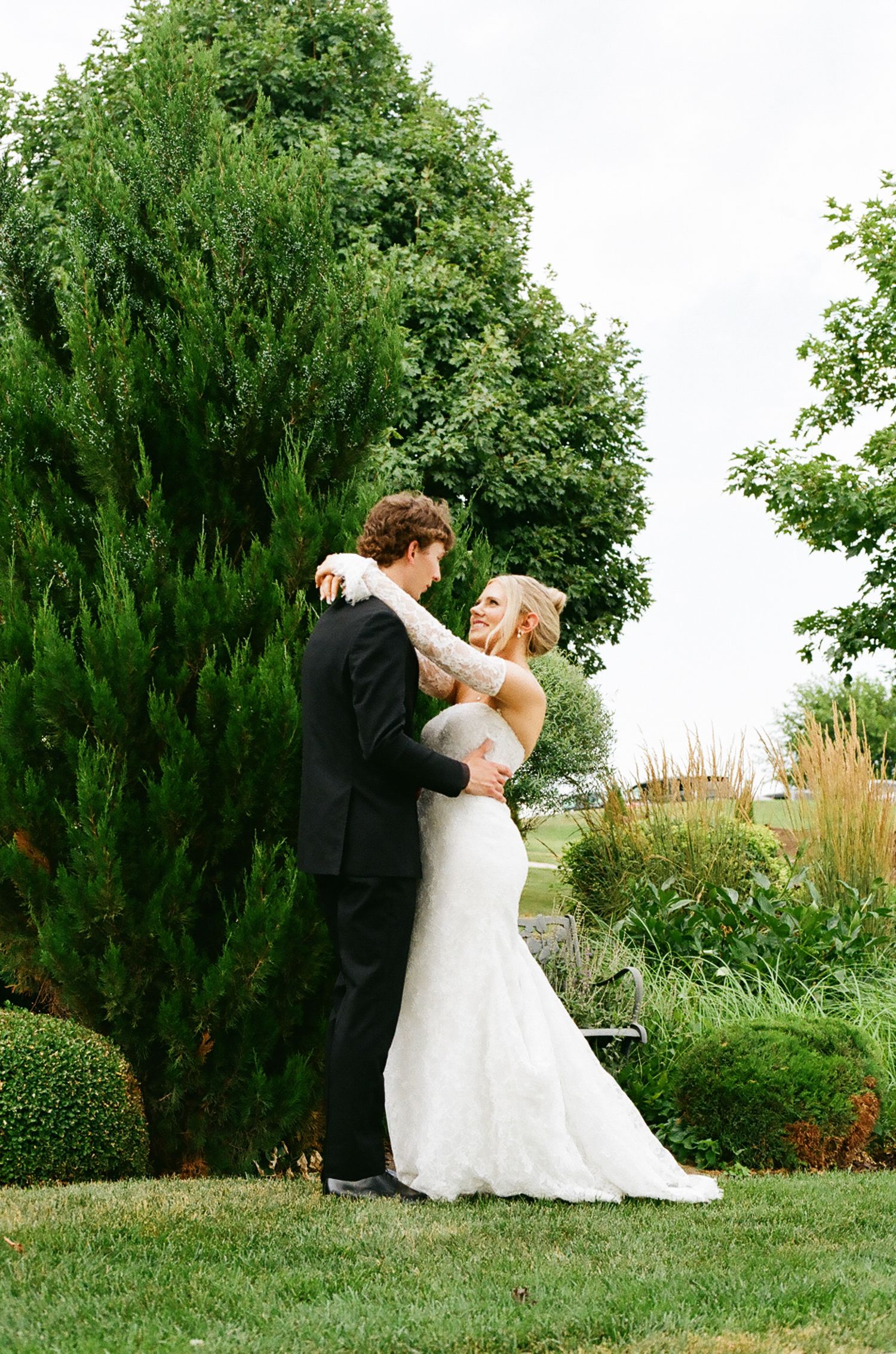 A bride and groom embrace in a lush garden, the bride wearing a fitted lace gown and the groom in a classic black tuxedo.