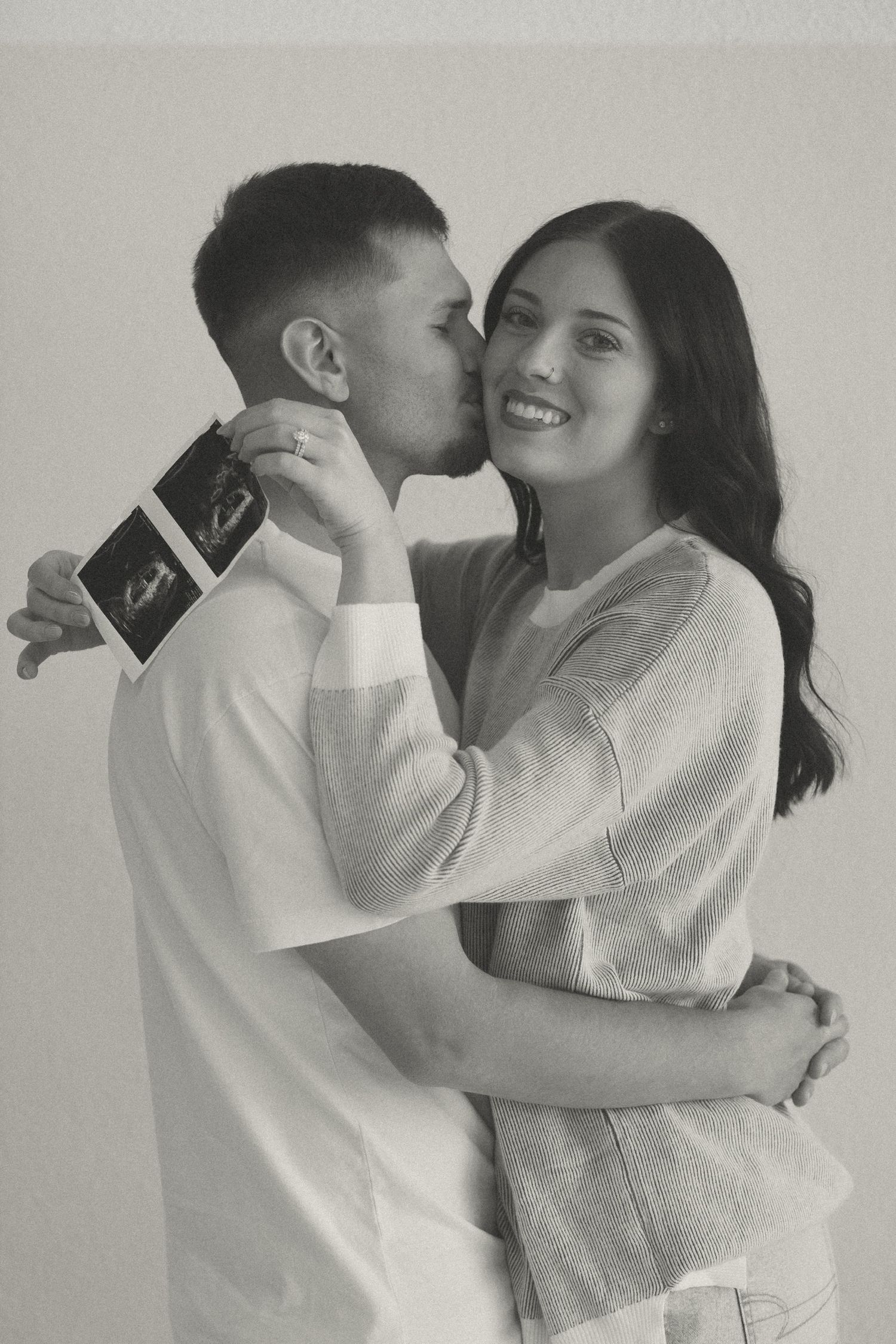 Black and white photo of man kissing woman on cheek while holding ultrasound photos.