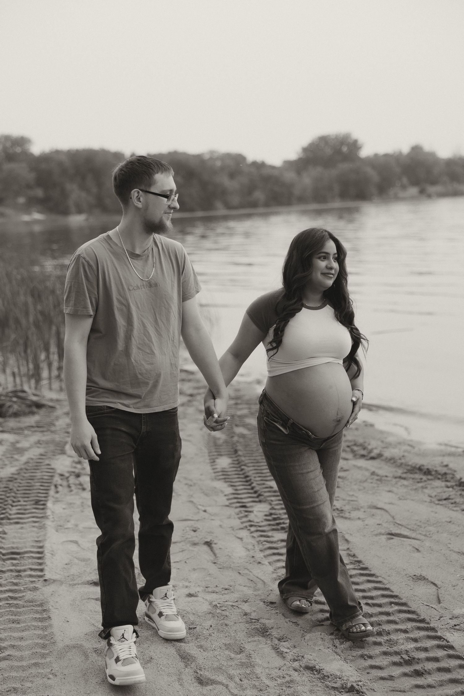 Black and white photo of pregnant couple holding hands walking along sandy riverbank.