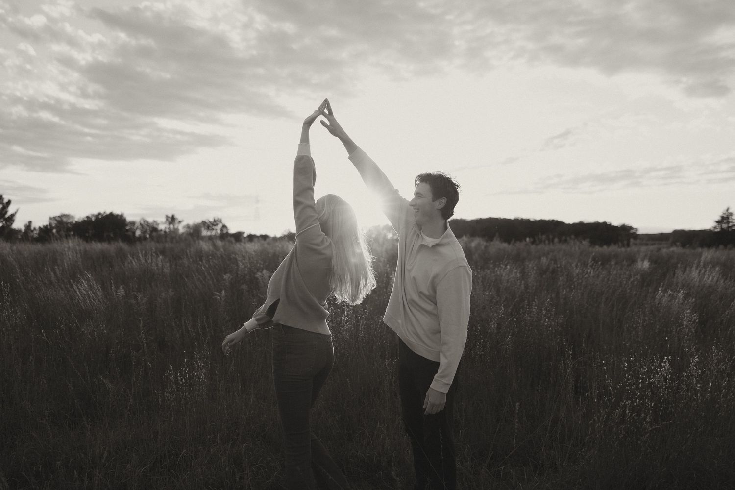 Black and white photo of a couple dancing and holding hands raised in a sunlit field.