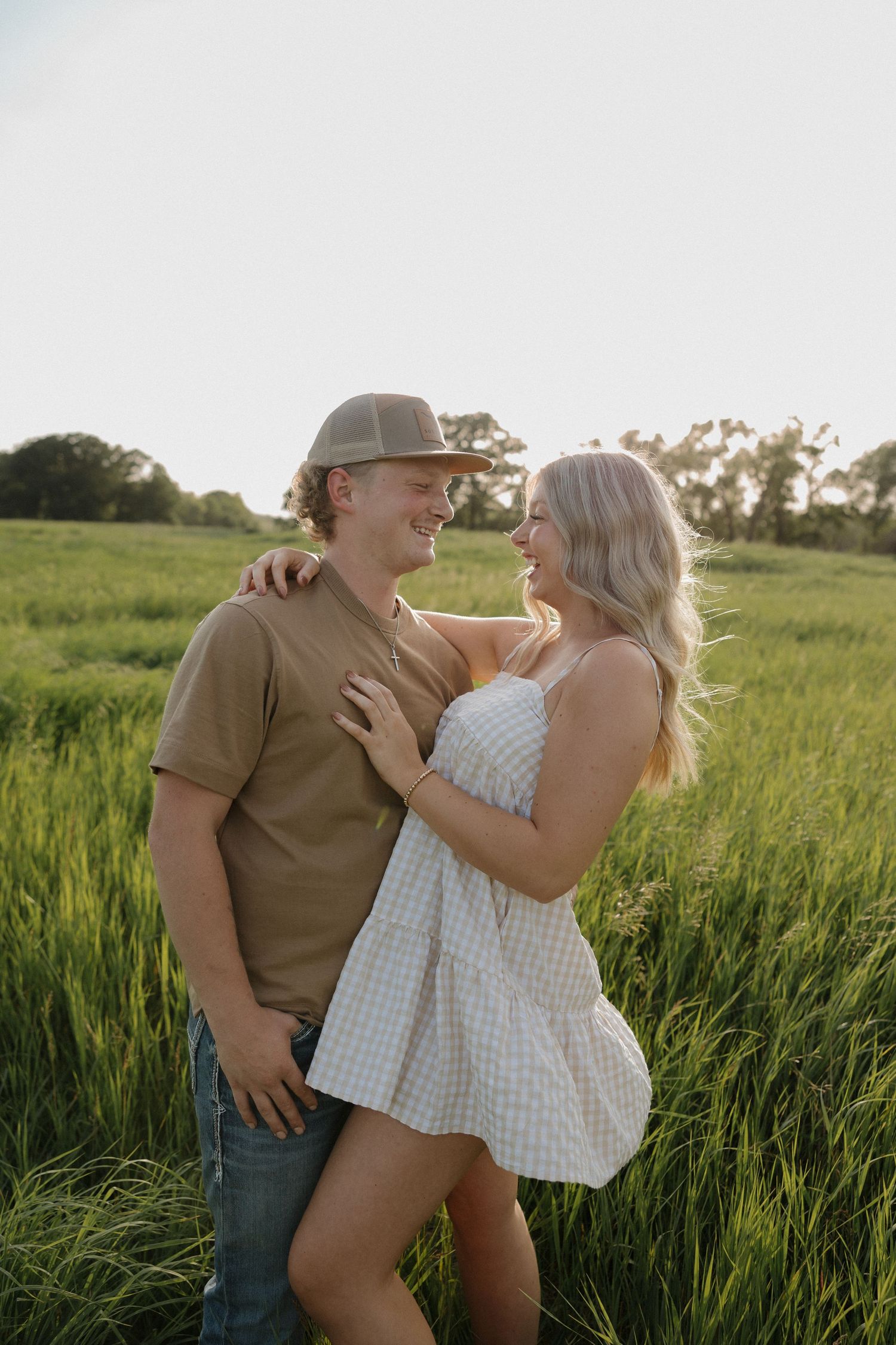 Smiling couple facing each other in sunlit green field, woman in white dress embracing man in cap.