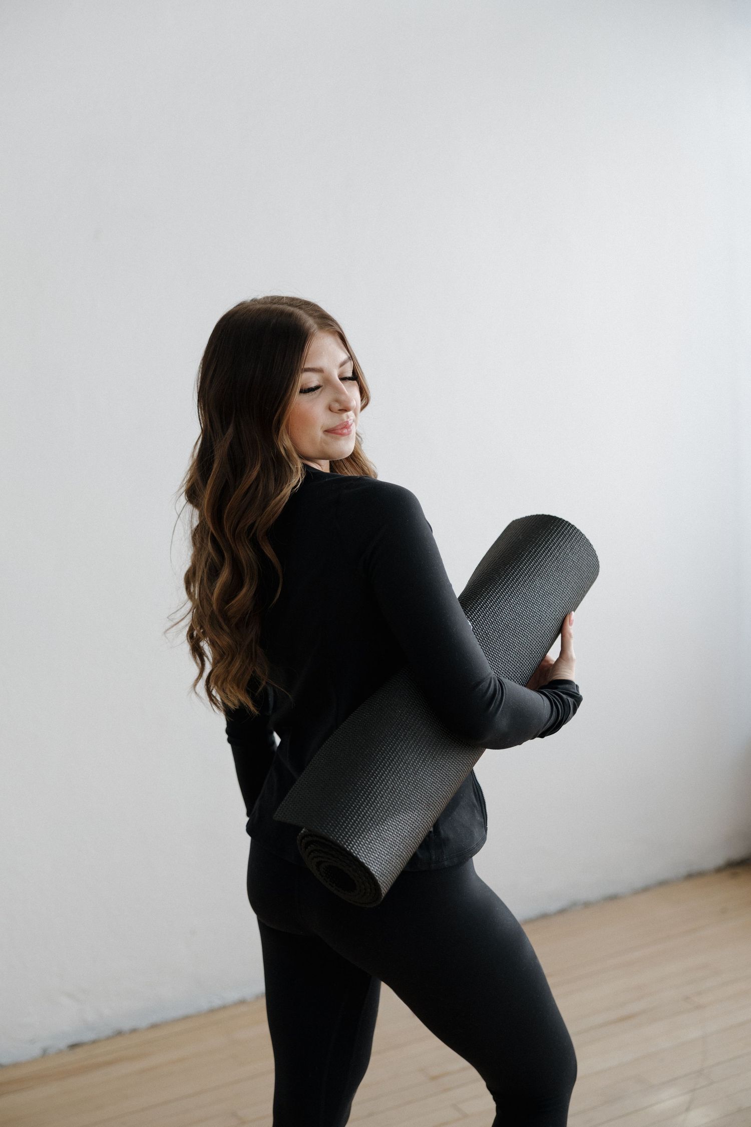Woman in black athletic wear holding a rolled yoga mat in a bright white studio space.